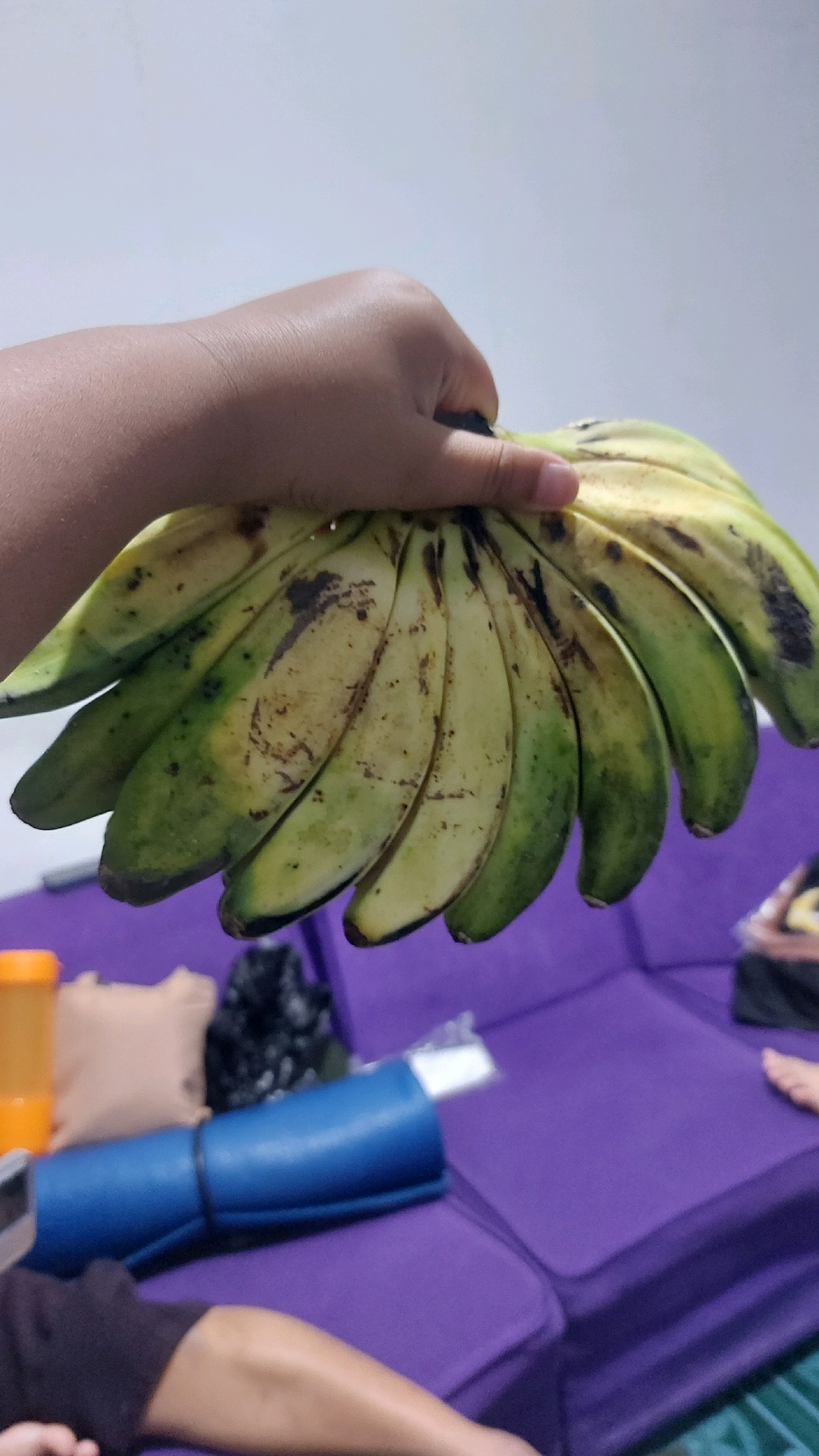 A bunch of green and yellow bananas, some with brown spots, are held by a person's hand against a light-colored wall. In the background, a purple couch is visible with various objects on it, including a blue rolled-up mat, an orange container, and a black object. Part of a person's leg and arm are also visible in the foreground, resting on the couch. The scene appears to be indoors, possibly in a residential setting in Salatiga, Indonesia.