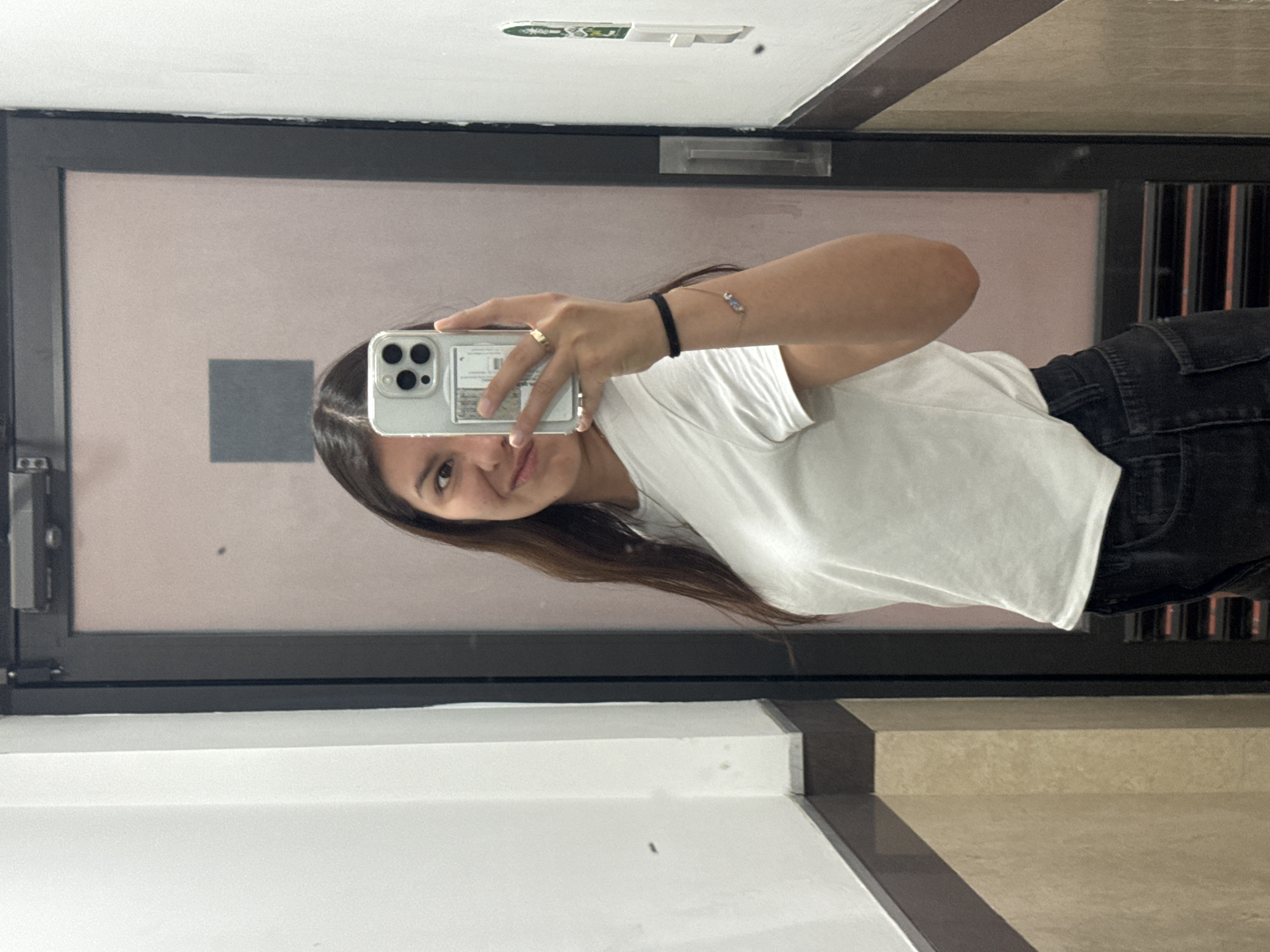 A woman takes a selfie in front of a mirrored door. The door, with a black frame, reflects a peach-colored wall. A gray square is visible on the wall within the mirror's reflection. The woman holds a smartphone in her right hand, her face partially obscured by the device. She wears a white t-shirt and dark pants. A gold ring is visible on her finger, along with two bracelets on her wrist. The floor is comprised of white and beige tiles. The location is San Andrés Cholula, Mexico.