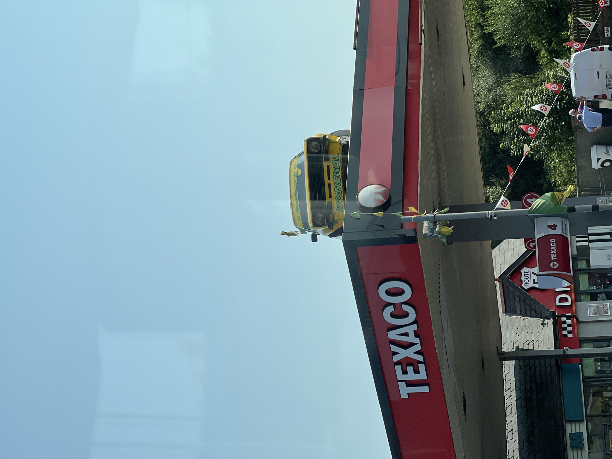 The image shows a Texaco gas station with a yellow car displayed on top of the canopy. The car has the word "DONEGAL" written on its front and "Good Luck" written on its windshield. The Texaco logo is prominently displayed on the red canopy. In the background, there are trees and a building with a "Route 53" sign. A white van is parked near the gas pumps, and two people are standing next to it. There are also several small flags strung across the area. The sky is clear and blue. The Texaco station is located in Letterkenny, Ireland.