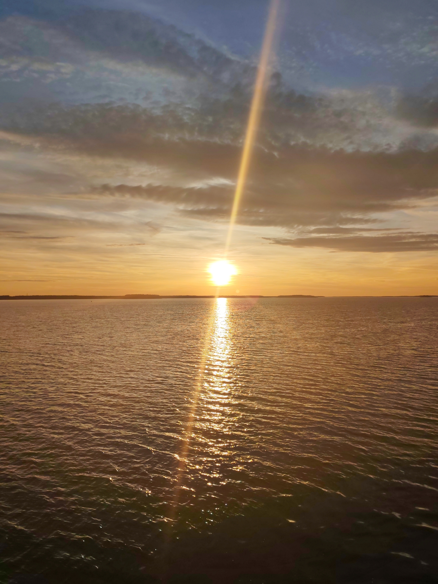 The image depicts a sunset over a body of water. The sun is positioned at the horizon, casting a bright, vertical beam of light upwards through the sky. Reflected sunlight creates a shimmering path on the water's surface, extending towards the foreground. The sky is filled with clouds, colored in hues of orange, yellow, and gray, indicative of the sunset. The water appears calm with gentle ripples. A dark strip of land is visible in the distance, possibly a coastline. The location and city are not discernable from the image.
