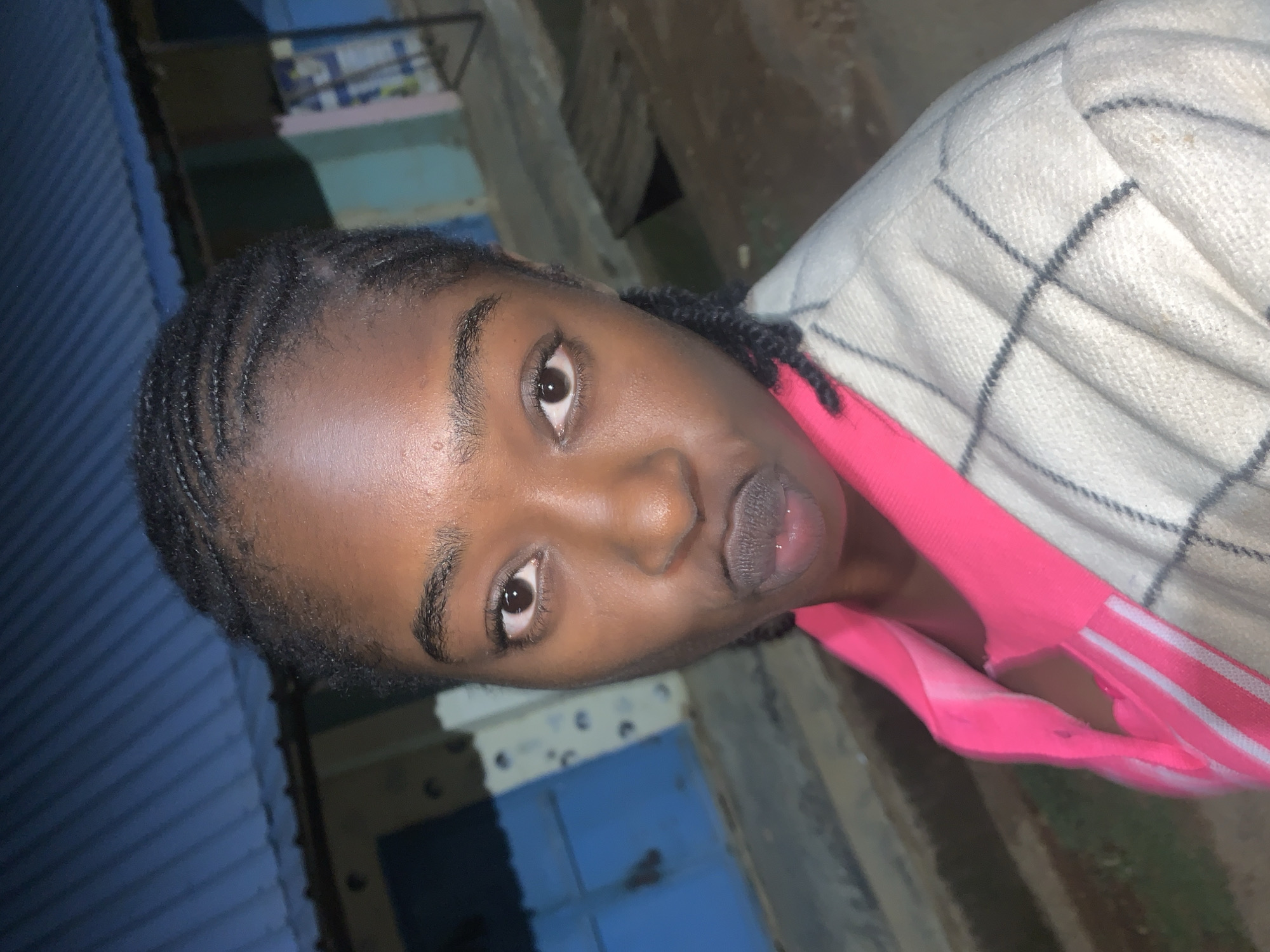 A young Black woman is depicted in a medium shot, looking directly at the camera with a direct gaze. Her medium-dark complexion exhibits a subtle sheen on the forehead and nose. Her hair is styled in cornrows pulled back from her face. Her eyebrows are dark and well-defined, and her eyes are dark brown. Her lips are slightly parted and puckered. She is wearing a bright pink collared shirt, with white horizontal stripes visible on the inner placket/collar. Over this, she wears an off-white or light gray jacket featuring a subtle dark plaid pattern composed of intersecting lines. The background, located in the city of Kabong, Nigeria, shows elements of an outdoor urban environment. To the left, a blue corrugated metal roof is prominent, below which a light blue building wall with irregular white markings is visible. To the right, another building section displays lighter blue, green, and a hint of pink coloration. The overall lighting appears to be dim or artificial, consistent with evening or night conditions.