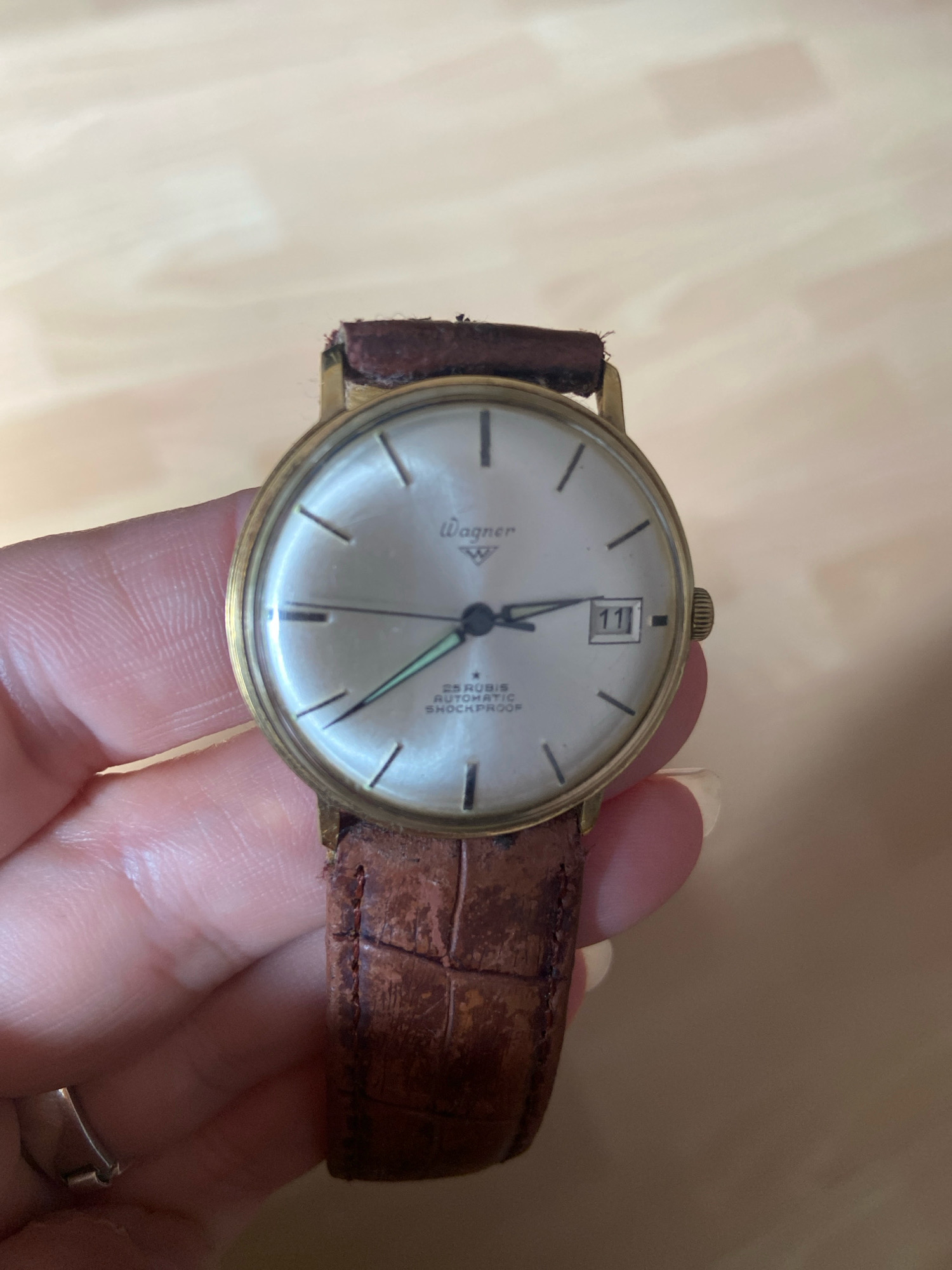 The image shows a close-up of a vintage Wagner automatic wristwatch with a brown leather strap. The watch face is light-colored with black hour markers and a date window showing "11". The watch is held by a person's hand, revealing skin texture and a ring on one finger. The background is a light-colored wooden surface, possibly a table or floor. The image appears to be taken indoors. There is no information in the image to indicate the location in the city of Kassel, Germany.