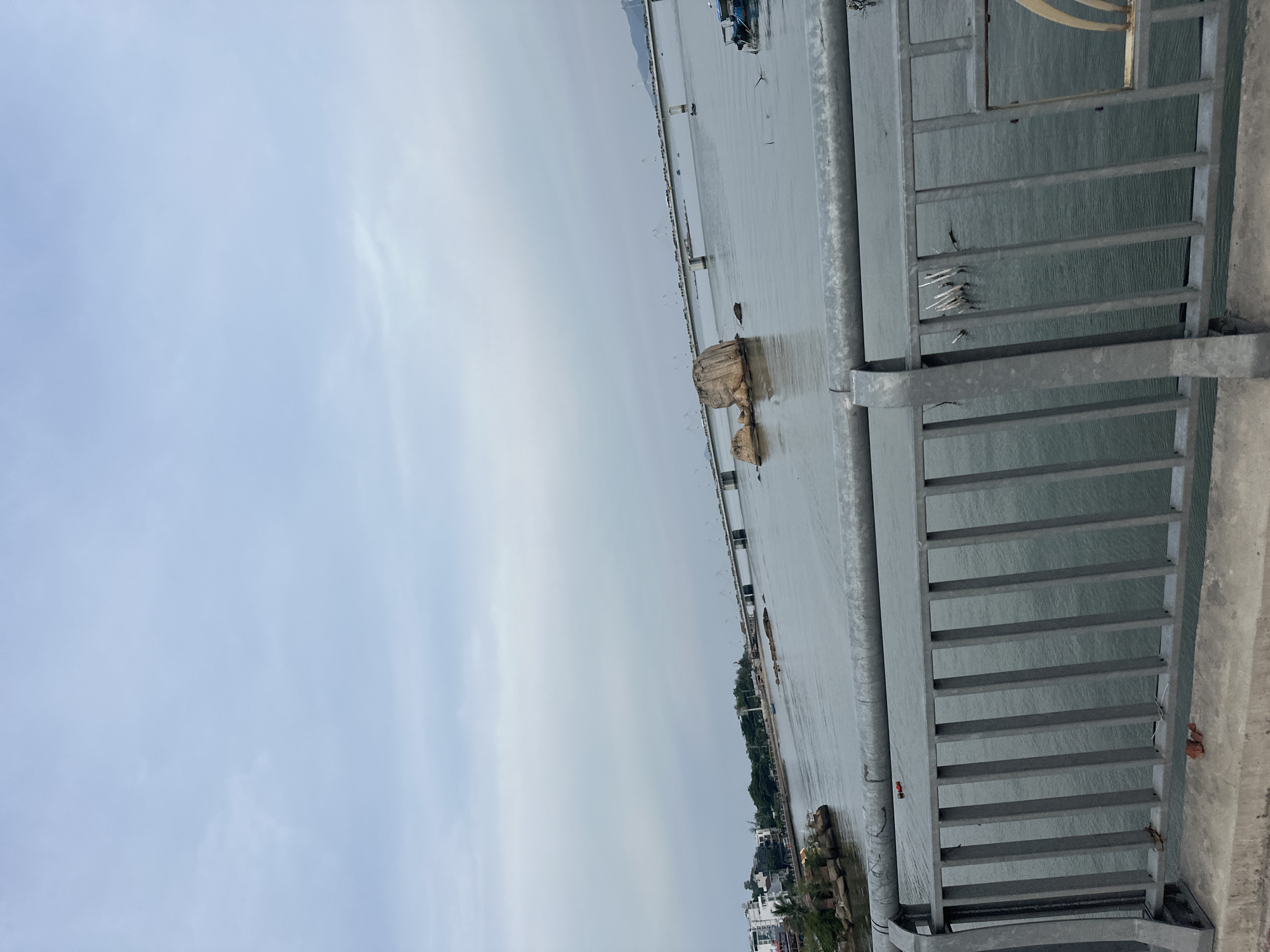 This is a wide shot of a body of water with a long bridge in the distance, viewed from a railing. The sky is overcast and pale blue, with some wispy clouds. In the foreground, a metal railing with vertical bars runs horizontally across the bottom of the frame. The railing is attached to a concrete base. The water is a dark gray, with small ripples. In the middle distance, there are several large rocks rising out of the water. A long bridge spans the water in the background, with cars and possibly some structures visible on it. To the left of the bridge, there is a shoreline with buildings and trees. A small blue boat is visible in the water to the right of the frame. There is no visible signage or identifying markers to confirm the location as Bac Nha Trang Ward, Vietnam.