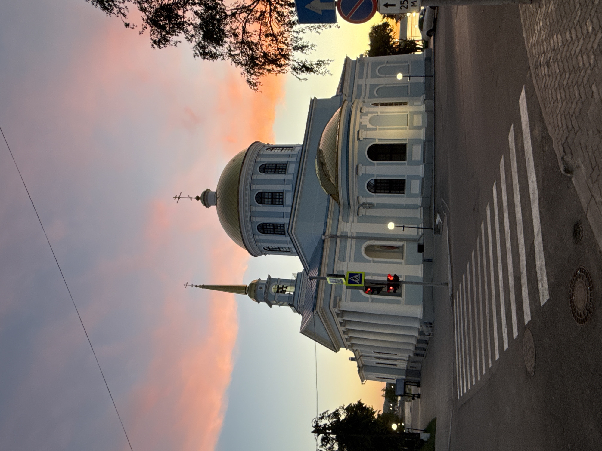 The image, originally rotated 90 degrees counter-clockwise, depicts a serene urban scene at either sunrise or sunset in Votkinsk, Russia.
The primary subject is a grand, light blue-grey building, likely a cathedral or large church, exhibiting classical architectural elements. It features multiple prominent golden domes and a tall golden spire topped with an Orthodox cross. The building's facade is adorned with columns, arched windows, and intricate cornices, illuminated by the soft, warm light of the sky. Several streetlights along the building's edge are illuminated, suggesting dusk or dawn.
In the foreground, a wide asphalt road stretches across the frame, featuring a clearly marked pedestrian crosswalk with white stripes. A red traffic light is visible on a pole beside the road, indicating a stop. The sidewalk adjacent to the building is paved with textured grey pavers, and two circular manhole covers are on the road surface.
The sky is a prominent feature, displaying a beautiful gradient of colors, transitioning from soft pinks, oranges, and yellows near the horizon to cooler purples and light blues higher up. This coloration strongly suggests the time of day is either sunrise or sunset. A dark, silhouetted tree with sparse foliage extends into the upper part of the frame, and a thin power line stretches across the sky.
Visible text and signs include: a blue rectangular road sign with an upward arrow; a red circular sign with a diagonal line (likely "No Entry" or "No Stopping"); a white rectangular sign with an upward arrow and "25 M"; and a yellow square sign with a black pedestrian symbol, indicating a pedestrian crossing. No people are visible, contributing to the tranquil atmosphere.