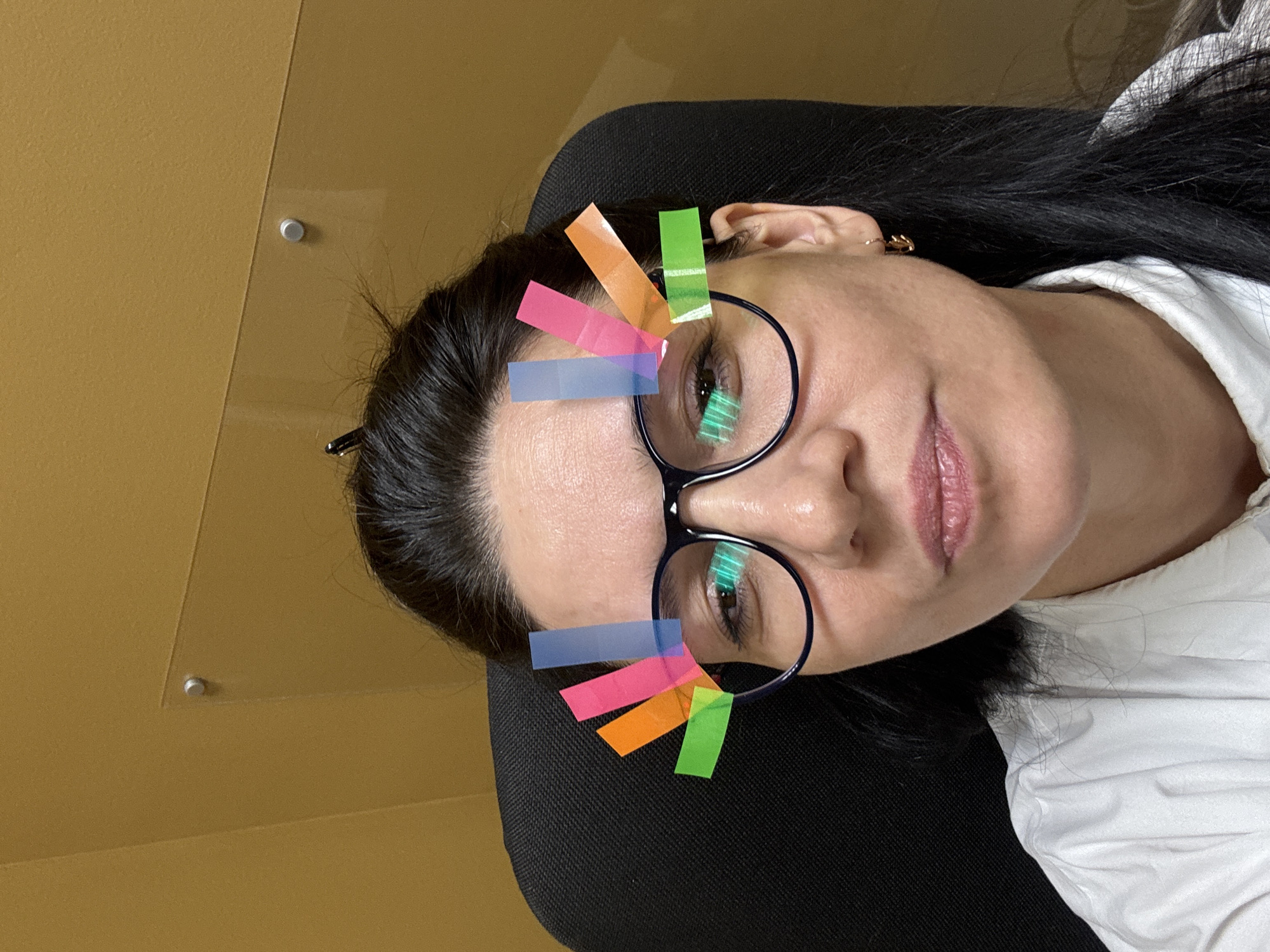 A woman with dark hair and glasses is lying in a black chair. Colored rectangular stickers are stuck on the glasses. She is wearing a white shirt. The background consists of a yellow wall. There is no information to verify if this image was taken in Chaikovsky, Russia.