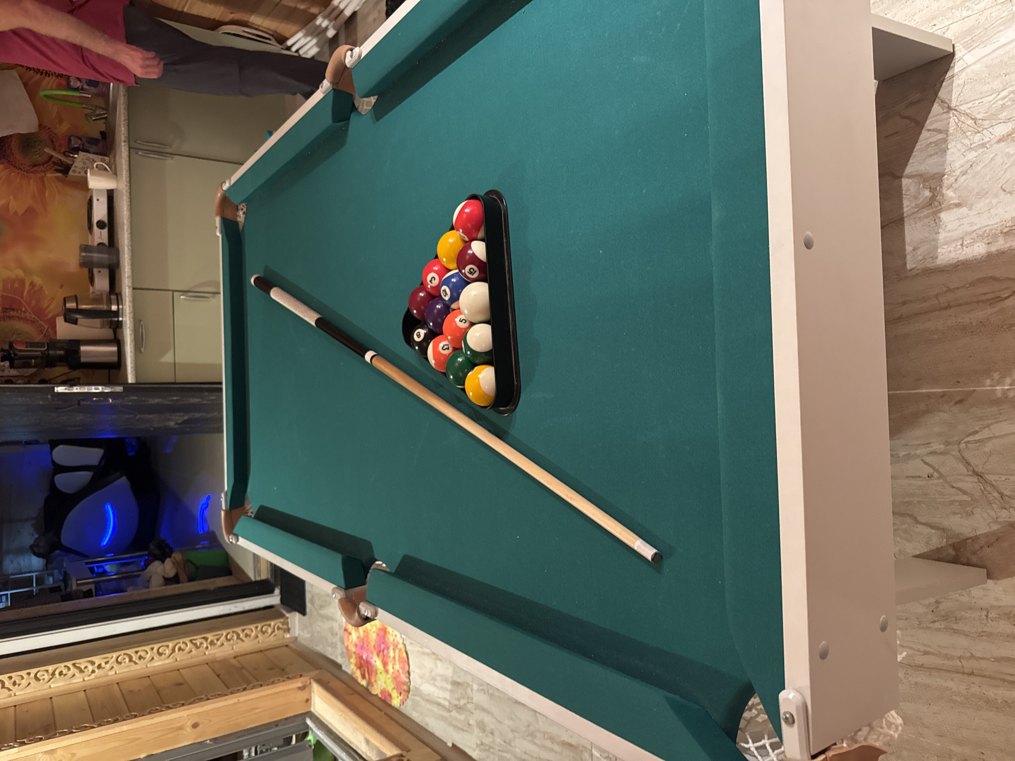 The image shows a pool table with balls racked and a cue stick resting on the felt. The pool table is green with white trim and is set up in a room with wooden walls and a tiled floor. In the background, there is a kitchen area with cabinets and a sink, and further in the background, there is a large inflatable object with blue lights, possibly a hot tub or a gaming pod. A person's legs and torso are visible on the right side of the image, wearing dark pants and a maroon shirt. The overall impression is a recreational space, possibly a basement or game room, with some of the amenities suggesting a domestic setting in Zelenaya Roshcha, Russia.