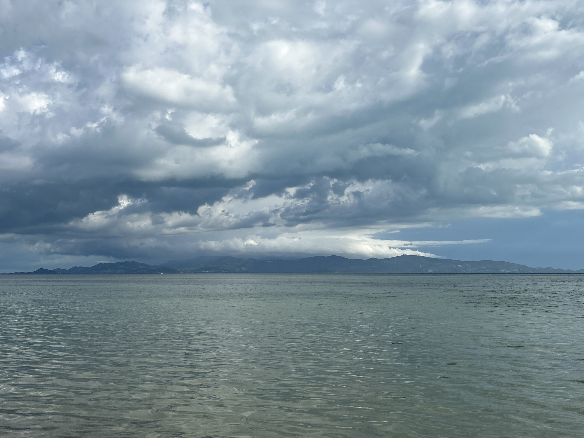The image depicts a body of water under a cloudy sky. The water is relatively calm, with subtle ripples reflecting the light. In the distance, a range of mountains or hills is visible on the horizon. The clouds dominate the upper portion of the frame, with varying shades of gray suggesting a potential storm or overcast weather conditions. The location is likely ตำบลเกาะพะงัน, Thailand.
