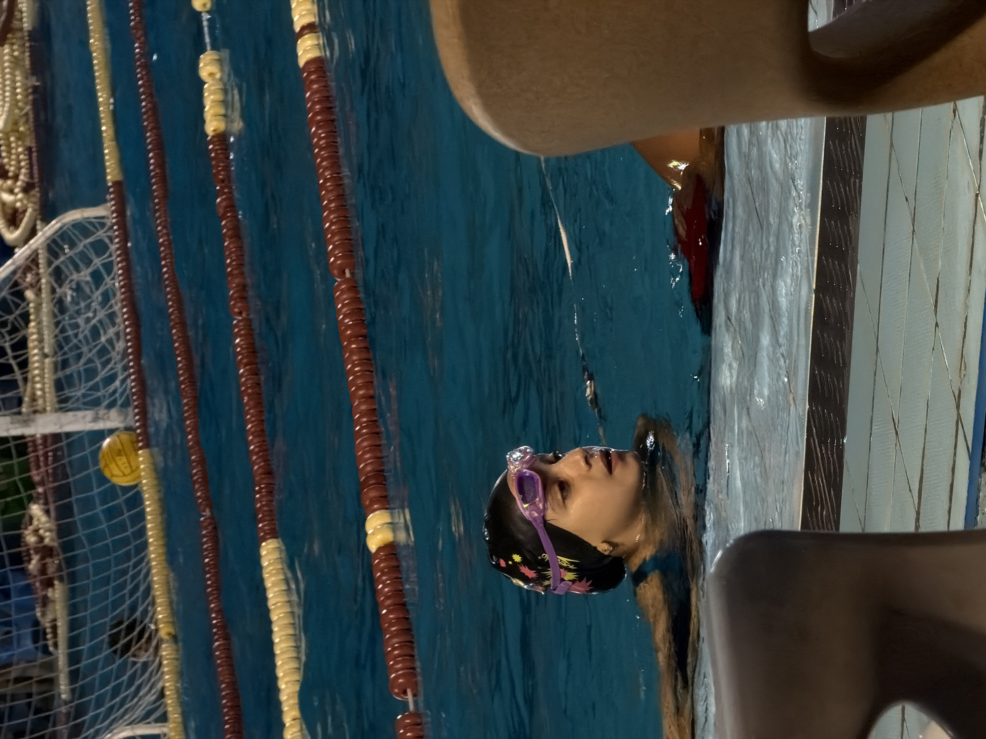 A young girl wearing a black swim cap with a floral pattern and purple goggles is in a swimming pool. She is looking to her right, with her mouth slightly open. In the background, there are lane markers in the pool, a water polo goal with a yellow ball, and a net. In the foreground, a person's shoulder and back are visible. The location is identified as a swimming pool in 6th of October City, Egypt.