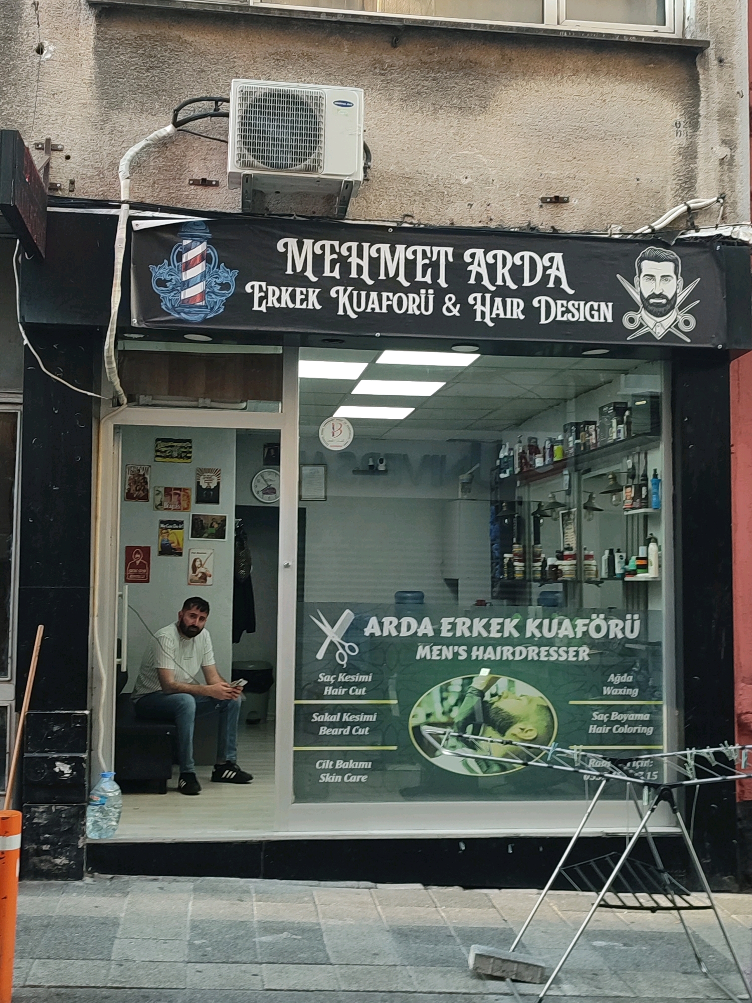 This is a street-level, eye-level, medium shot of a barber shop in Kadıköy, Turkey. The shop's facade is black, with a large banner above the entrance that reads "MEHMET ARDA ERKEK KUAFFÖRÜ & HAIR DESIGN." The banner features a red, white, and blue barber pole graphic on the left and a stylized illustration of a man with scissors on the right. The shop's interior is visible through the open doorway and large glass window. Inside, shelves are stocked with various hair care products. A man, appearing to be an employee, is seated inside the shop near the entrance, looking towards the camera. He is wearing a striped t-shirt and blue jeans. Behind him, the shop walls are decorated with framed posters and a clock. Outside the shop, a clothes drying rack with some items on it stands on the sidewalk. The street in front of the shop is paved with gray and white square tiles. An air conditioning unit is mounted on the wall above the shop entrance.
