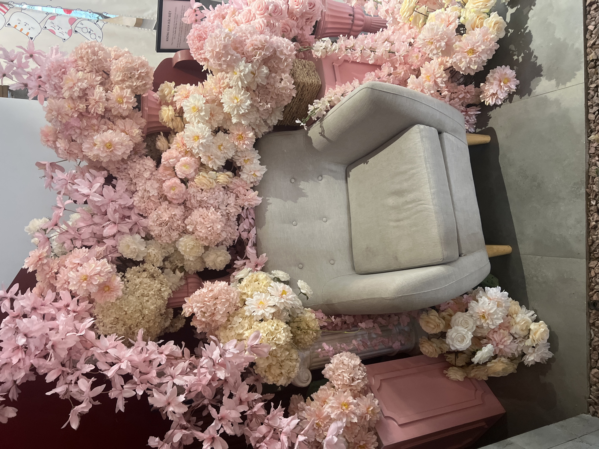 This image captures a vibrant and heavily decorated indoor or semi-enclosed space, likely a lounge area or photo booth, within a commercial establishment in Quy Nhon Ward, Vietnam. The scene is dominated by an abundance of artificial flowers in shades of pink, cream, and pale yellow, creating a lush, romantic ambiance.
A light grey upholstered armchair, featuring tufted buttons on its backrest and light wooden legs, is centrally positioned and surrounded by the extensive floral arrangements. The flowers include large clusters of pom-pom-like blooms resembling chrysanthemums or hydrangeas, delicate branches of cherry blossom-style flowers, and bouquets of roses and other rounded blossoms. These are artfully arranged around the chair, on the floor, and on several pink decorative boxes or pedestals, one with an ornate frame design. A pink fluted vase and a woven basket are also nestled amongst the flora.
The flooring consists of grey tiles or concrete, upon which strong shadows are cast by the flowers and the armchair, indicating bright artificial lighting. On the far right, a strip of reddish-brown pebbles suggests a transition to an outdoor or semi-outdoor area.
In the top left corner, a reflection in what appears to be a mirror reveals cartoon cat drawings with speech bubbles. A black framed sign, partially obscured by flowers, is visible near the top center. It displays the Vietnamese text "KHOANG GIAN DAC BIET," which translates to "SPECIAL SPACE."
The scene is devoid of people or explicit activity, serving as an aesthetically pleasing and inviting backdrop. The soft colors and profusion of flowers evoke feelings of warmth, tranquility, and aesthetic appeal. The visible Vietnamese text reinforces the provided location context of Quy Nhon Ward, Vietnam.