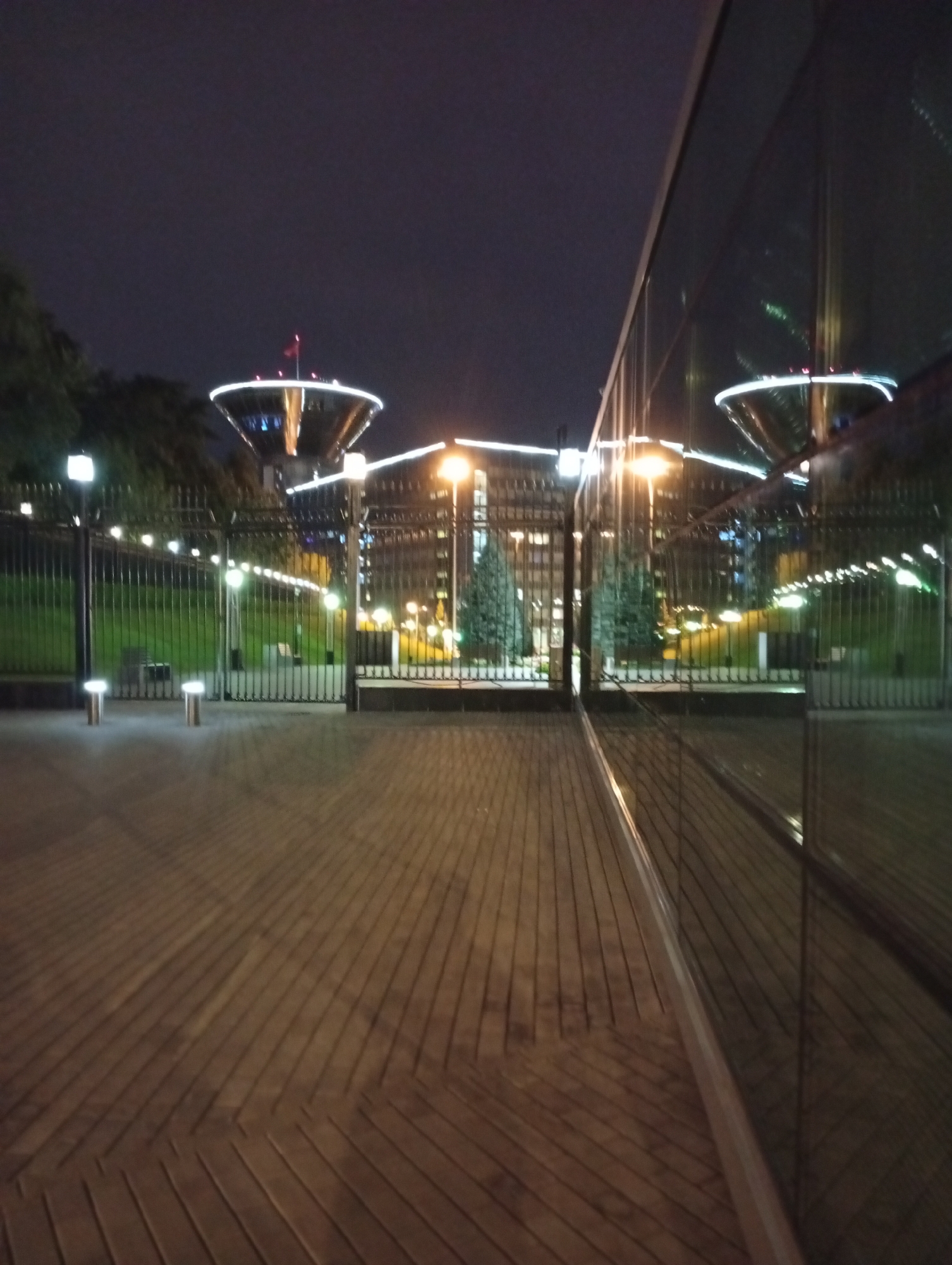An outdoor, eye-level, medium shot captured at night shows a paved walkway with distinct wood-patterned tiling. To the right, a large, reflective glass wall dominates the frame, mirroring the illuminated structures across the walkway. Beyond the walkway, a black metal fence encloses a grassy area with trees and illuminated light posts. In the background, two large, futuristic-looking structures with white illuminated rims and red flags on top are visible, suggesting a public or architectural landmark. The sky is dark and appears to be overcast. The overall scene conveys an urban nighttime ambiance.