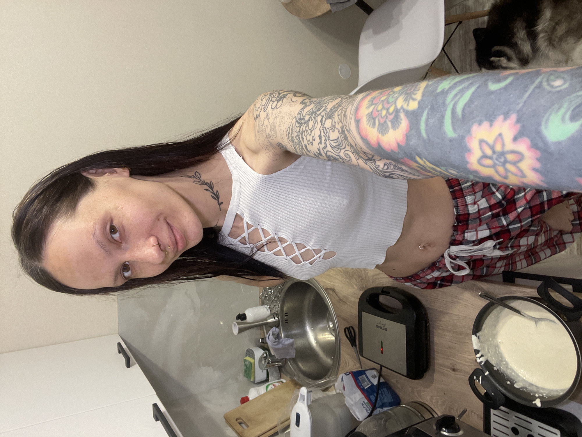 An indoor, slightly tilted, eye-level shot captures a young woman in a kitchen setting, likely taking a selfie. She has long, dark hair, dark eyes, and a neutral expression with a slight smile. She has a gold hoop nose ring and another stud on her nose. A small, delicate leaf-patterned tattoo is visible on her neck. Her left arm and shoulder are covered in intricate black and white tattoo designs, while her right arm (closer to the viewer) features extensive colorful floral tattoos in shades of pink, yellow, and green on a dark background. She wears a white ribbed crop top with a distinctive criss-cross lace-up detail on the front and red and white plaid shorts. A belly button piercing is also visible.
The woman is standing in what appears to be a modern kitchen. To her left, a stainless steel sink is visible with a faucet, sponge, and various kitchen items and bottles, including a wooden cutting board. On the wooden countertop to her right, a black and silver sandwich maker with the brand name "STINE" and a red indicator light is present, alongside a black pot filled with a light-colored batter (possibly for pancakes or waffles) and a spoon. In the background, behind a white chair, a fluffy dog with dark and light fur is partially visible. The walls are light-colored, and there are white and gray cabinets with black handles. The scene suggests she is engaged in or pausing during food preparation. The location context provided is Izhevsk, Russia.