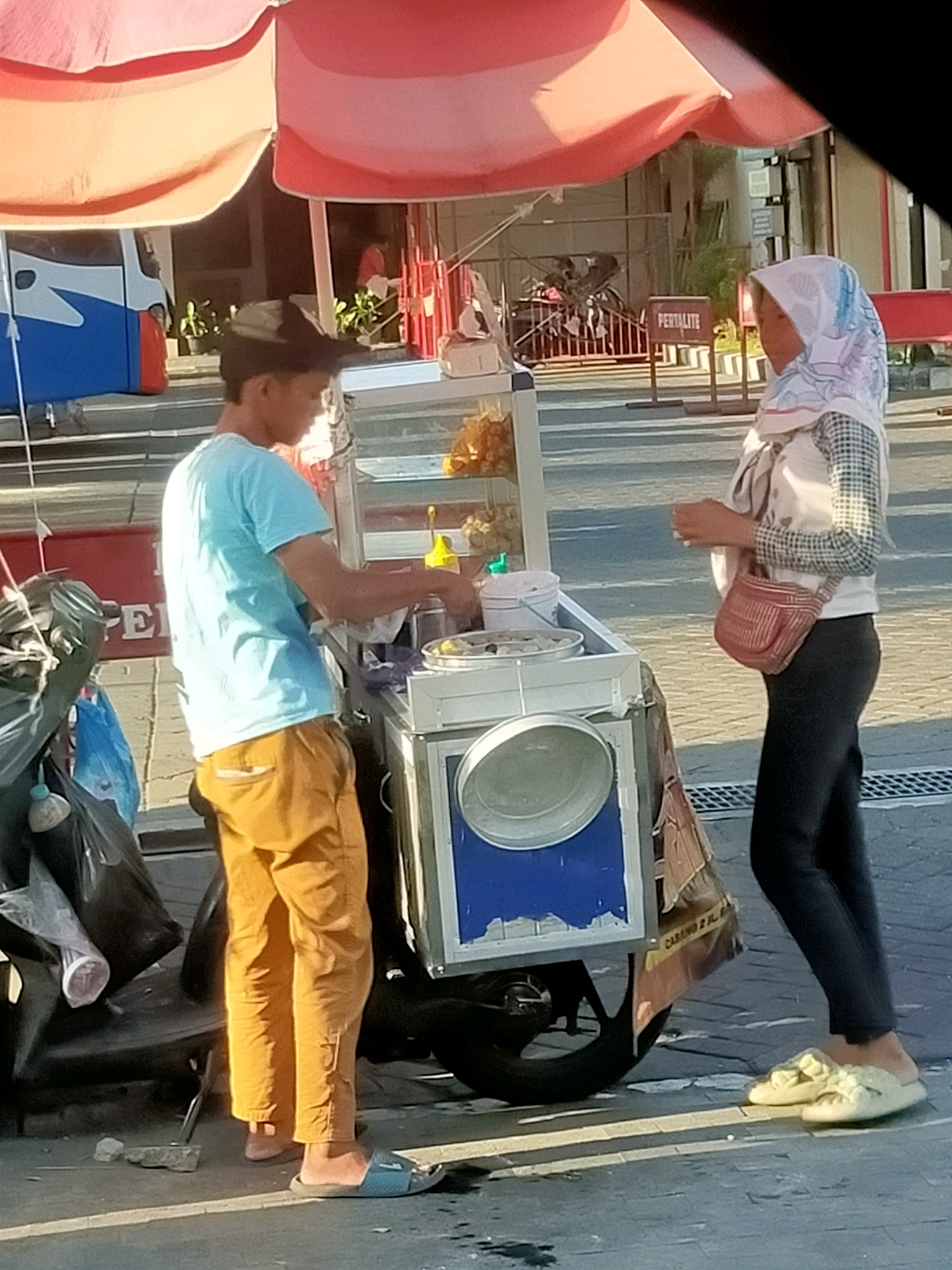The image depicts a street scene in Surabaya, Indonesia, featuring two individuals standing next to a food cart. A man, wearing a light blue t-shirt and orange pants, is behind the cart, seemingly preparing food. A woman, dressed in a headscarf and a checkered shirt over a light-colored vest, stands in front of the cart, holding a small red bag. The food cart itself is equipped with a display case containing various food items and containers. In the background, there is a bus, a fence with a "PERTALITE" sign, and other elements of an urban environment. The ground appears to be paved with cobblestones and a road. The overall lighting suggests it is daytime, with sunlight casting shadows.