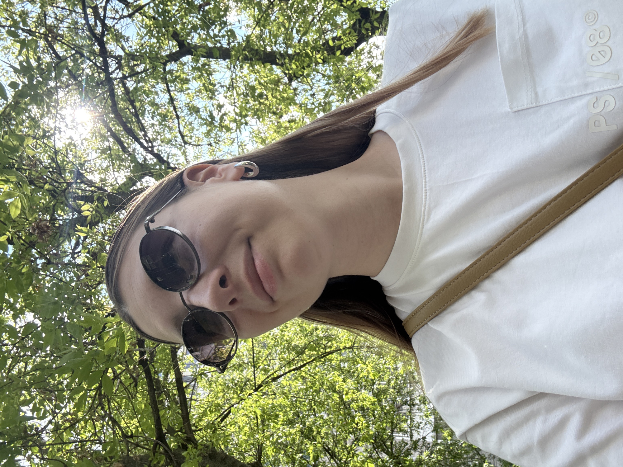 A woman with long brown hair, wearing a white t-shirt, sunglasses, and earrings, is seen from a low angle against a backdrop of green trees and a partially visible blue sky. Sunlight filters through the leaves. The t-shirt has the letters "PS/88" visible on the front. Based on the style of trees and the light, the location is possibly in a park or green space. The image does not provide enough information to confirm the location in Moscow, Russia.