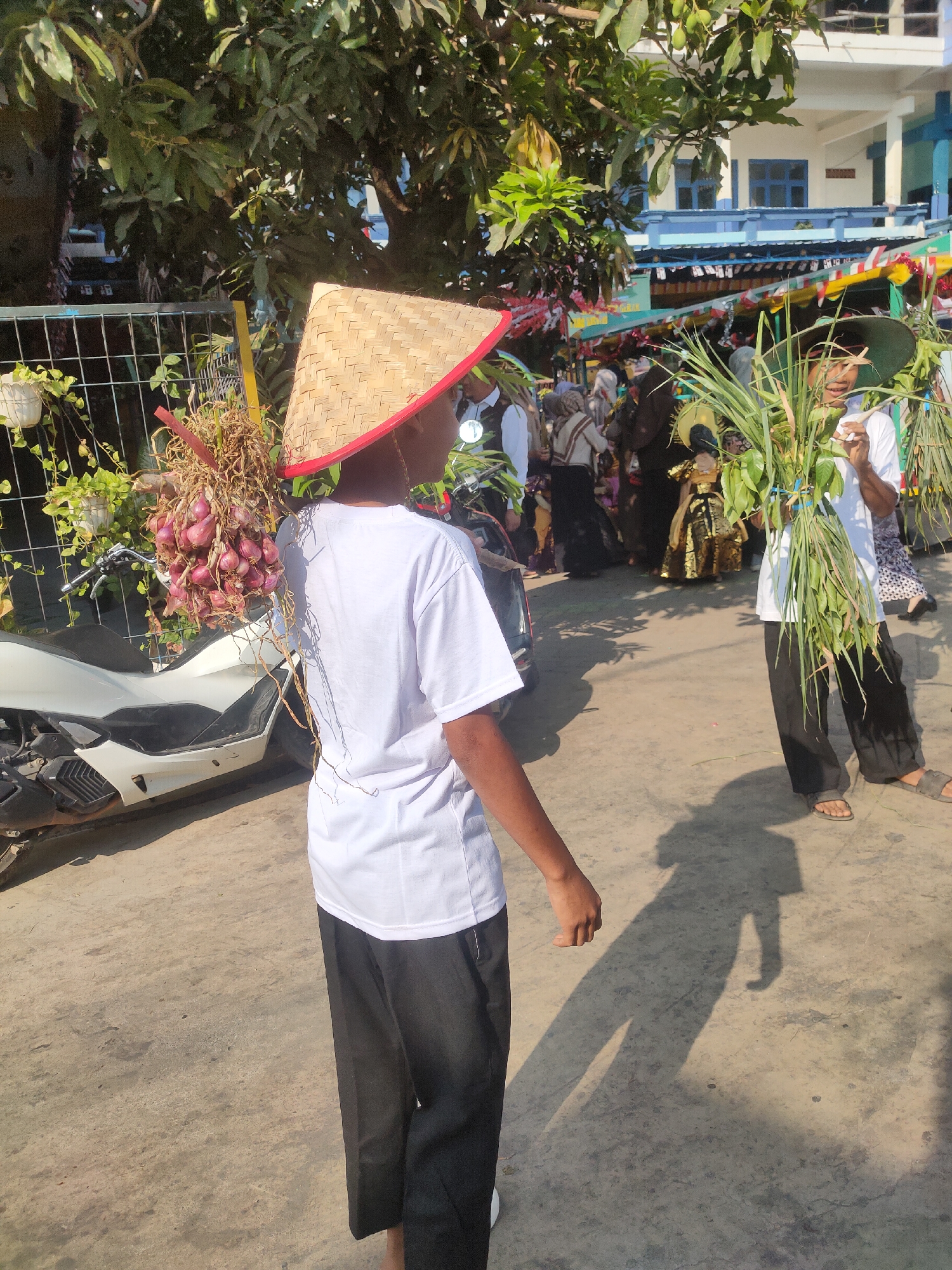 In Payaman, Indonesia, a young person wearing a conical straw hat and a white t-shirt carries a large bunch of red onions on their shoulder, their back facing the camera. They are standing next to a white motorcycle. Another individual, also wearing a white t-shirt and black pants, stands to the right, holding a large bundle of green stalks, likely lemongrass or similar. In the background, a crowd of people are visible, some in traditional attire, and a building decorated with colorful banners suggests a festive event or celebration. Trees with green leaves provide shade, and the ground is paved with concrete. The scene appears to be a street during daylight hours.