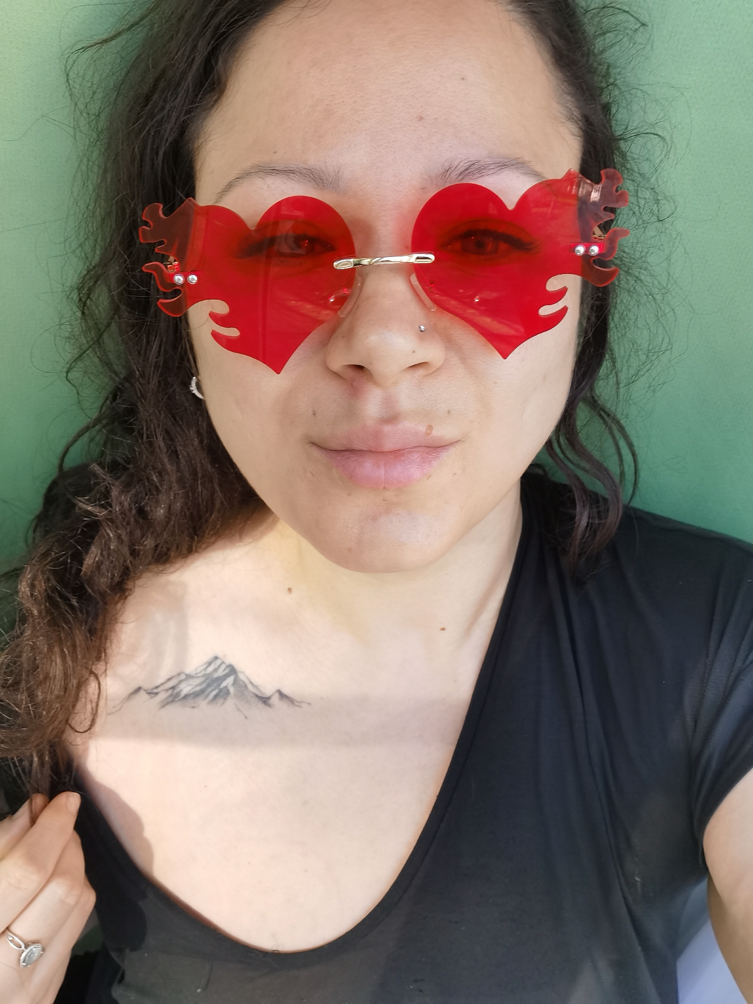 A medium close-up shot shows a woman wearing heart-shaped red sunglasses with flame-like detailing at the edges. She has dark, curly hair and a nose piercing. She is wearing a black v-neck shirt, and a tattoo of a mountain range is visible on her chest. The background is a solid green color. The location is not discernible from the image.