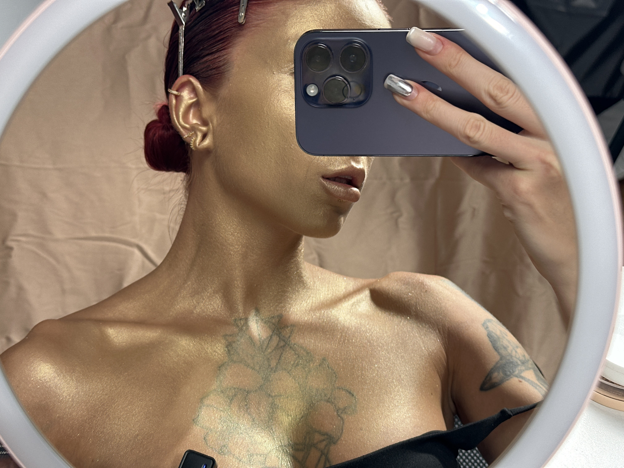 This media file is a mirror selfie featuring an individual, likely female, with a striking appearance, taken in what appears to be an indoor setting. The mirror itself is round and has an illuminated ring light, casting a bright glow and creating soft reflections. The background reflected in the mirror is a light, draped fabric, possibly a beige or light brown sheet or curtain.
The subject's skin, particularly on her face, neck, chest, and shoulders, is covered in a shimmering, golden body paint or highlight, giving her a metallic and radiant look. Her hair is a dark red or auburn color, styled back into a neat, low bun, with several silver hair clips visible near her scalp, suggesting hair preparation or styling. She wears multiple small, gold-colored ear cuffs or earrings on her visible left ear.
She is holding a dark-colored smartphone, likely an iPhone due to the distinct camera array, in her right hand to take the selfie. The phone covers the upper part of her face, with only her lips, which are slightly parted and appear to have a glossy finish, and chin visible. Her fingernails are manicured, with the thumb nail featuring a clear or light polish, and the index and middle fingers adorned with a reflective, metallic silver polish.
The subject is wearing a black top or bra strap, visible at the bottom of the frame. She has several visible tattoos: a large, intricate, possibly floral or geometric design on her upper chest/sternum area, and another tattoo on her left upper arm/shoulder, appearing to be an animal or creature.
The activity depicted is the act of taking a mirror selfie, possibly as part of a styling or preparation process for an event or photoshoot. While the location context is given as North Kuta, Indonesia, there are no specific visual cues within the image to confirm this geographic location. No discernible text is visible in the image.