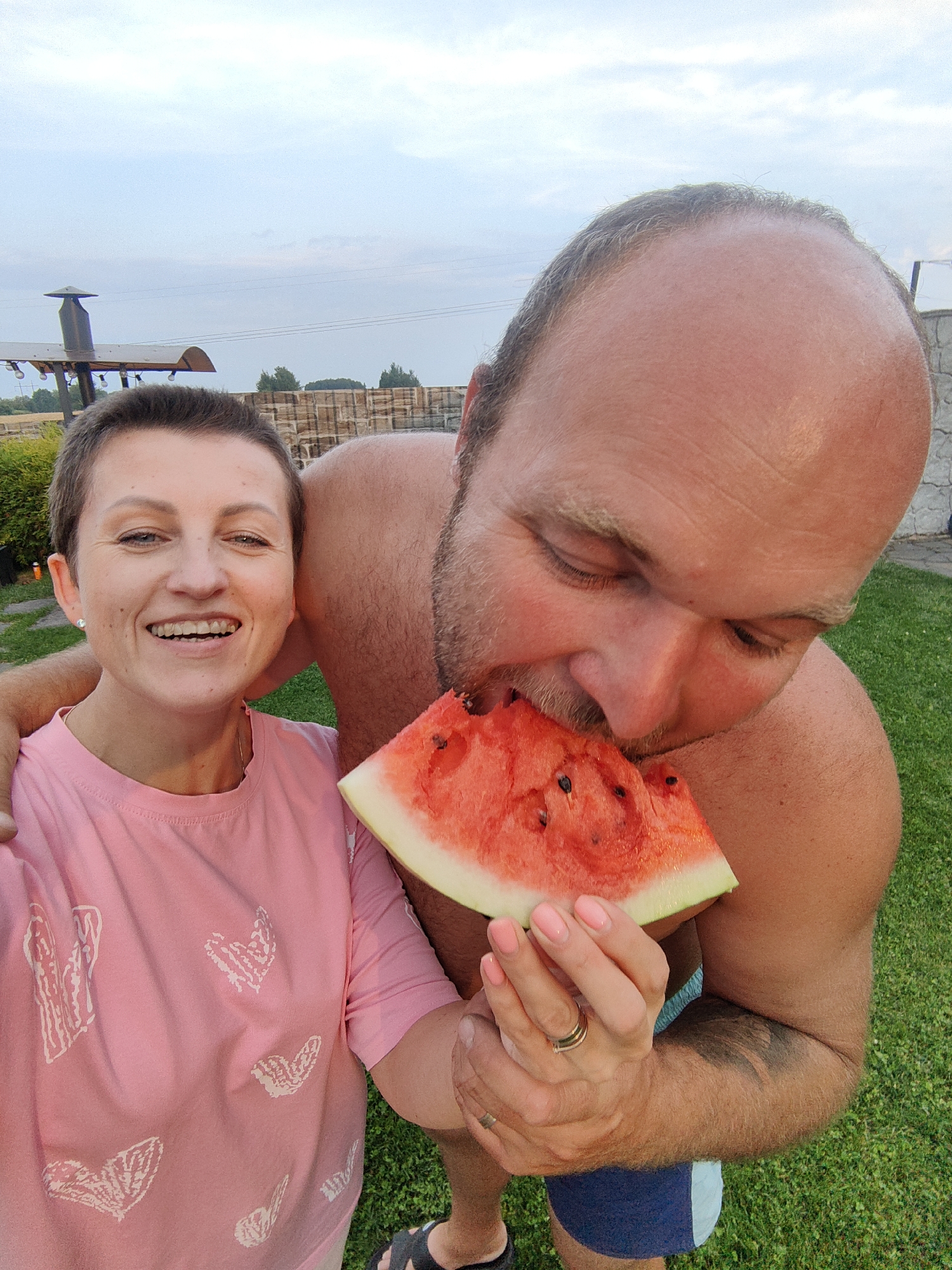 In a residential backyard in Фаніпальскі сельскі Савет, Belarus, a couple is sharing a slice of watermelon. The woman, with short brown hair and a pink t-shirt adorned with white heart designs, smiles broadly as the man, whose head is tilted down as he takes a bite from the watermelon, has his arm around her. The man is shirtless, and the woman is wearing a pink t-shirt. The scene unfolds on a green lawn, with a stone wall and some foliage visible in the background under a cloudy sky.