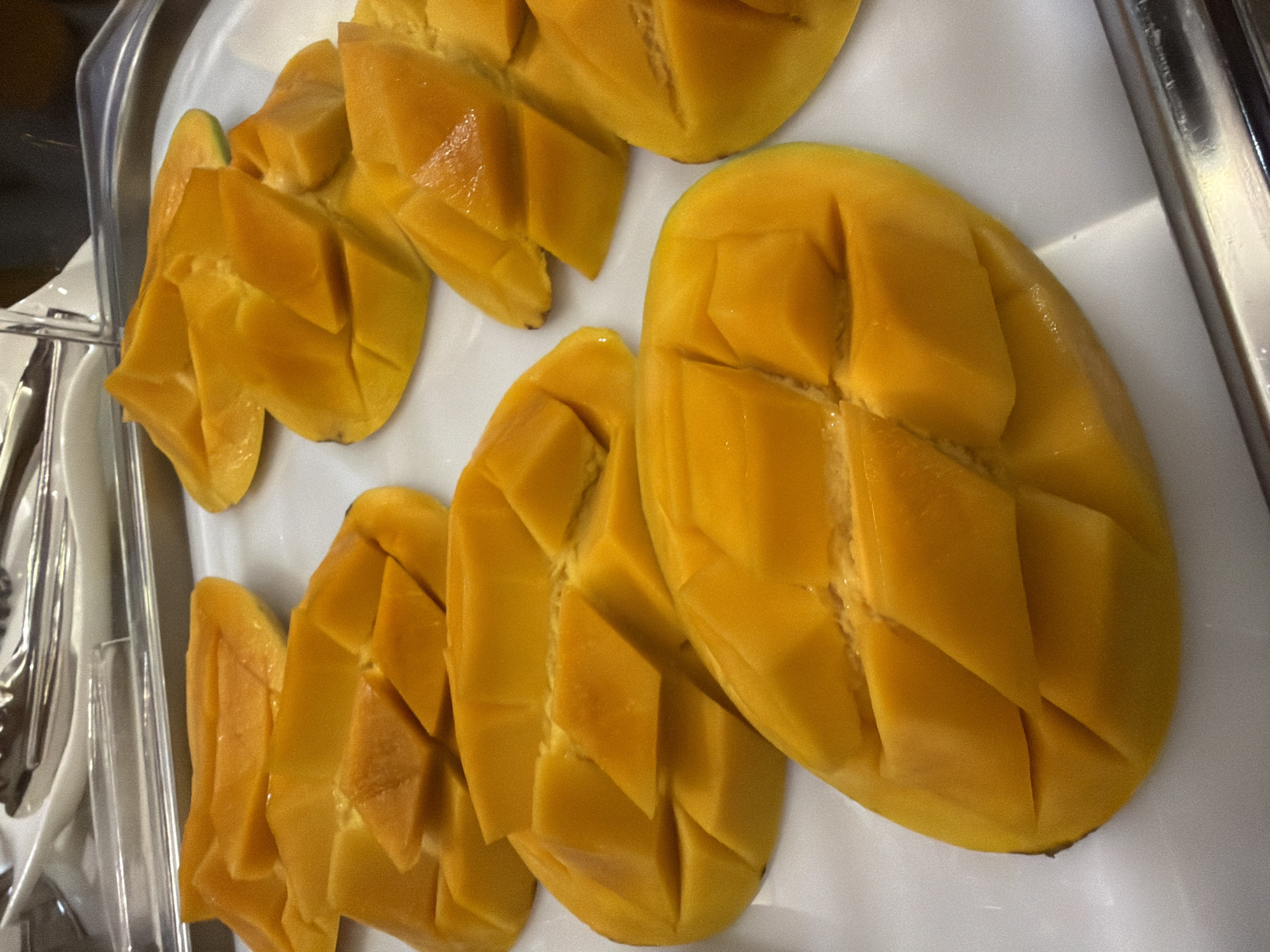 The image displays several halved and cubed mangoes arranged on a white tray. The mangoes are a vibrant yellow-orange color, with the flesh diced in a crosshatch pattern. The tray is placed on a stainless steel surface, and in the background, there are tongs and a white dish, suggesting a buffet or serving setting. The location is not discernible from the image.