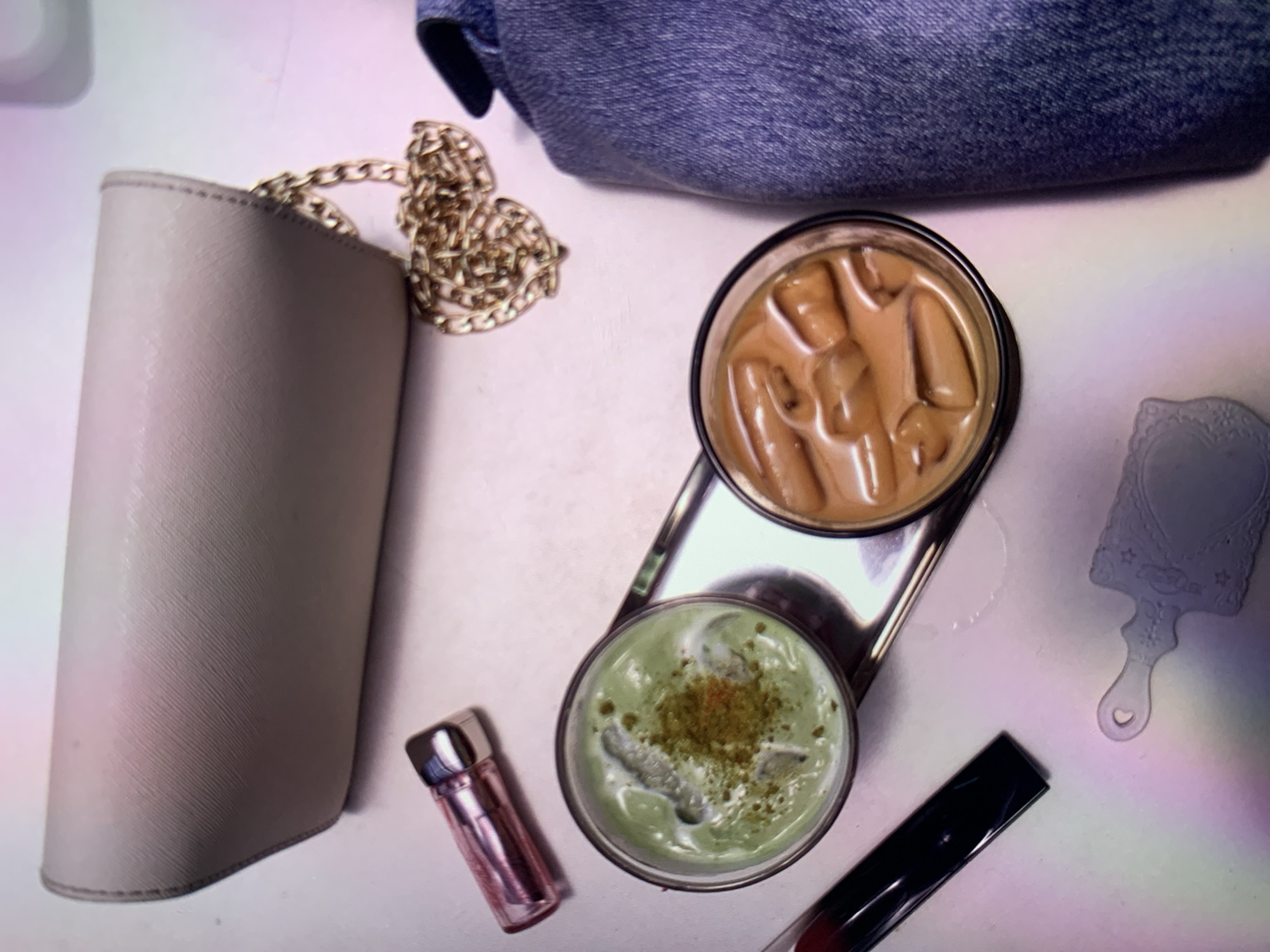 The image depicts a flat lay arrangement of several items on a white, possibly textured surface. In the upper portion of the image, a light-colored clutch purse with a gold chain is visible. To the left of the purse, a pink metallic lipstick or similar cosmetic tube is placed. Below these items, on a silver tray, are two glasses of iced beverages. The glass on the left contains a light green drink, likely matcha, with ice cubes and a dusting of green powder on top. The glass on the right contains a brown beverage, possibly iced coffee or tea, with ice cubes. To the left of the glasses is a dark-colored object, possibly another lipstick or pen. In the lower center of the image, a small, decorative, light-colored item with a patterned edge and a heart-shaped cutout is present. To the right of the glasses, a portion of dark blue denim fabric, likely from jeans or a jacket, is visible. The lighting in the image creates a slightly blurred and artistic effect with visible light flares. There is no discernible location in the city of Ciamis, Indonesia.