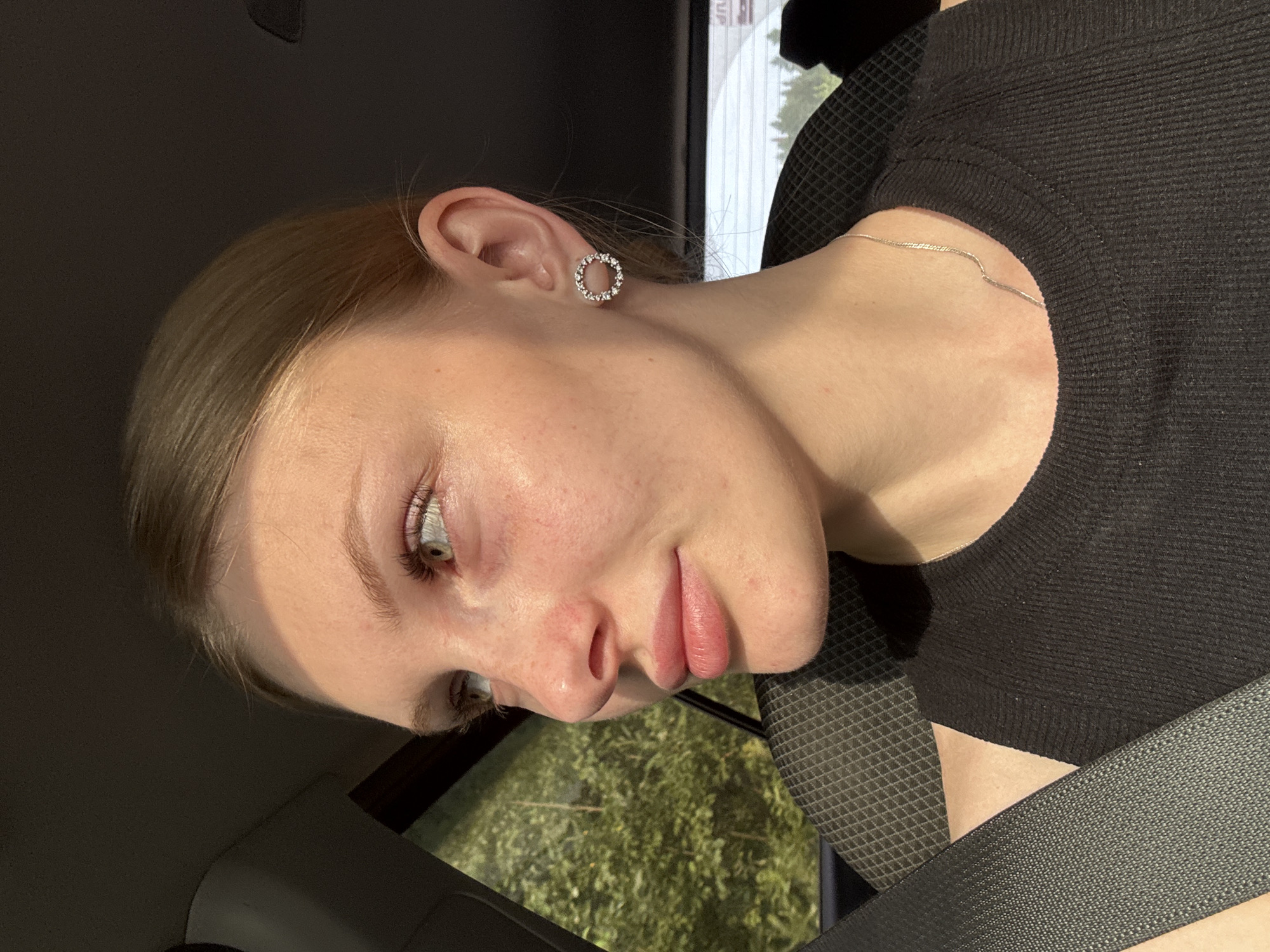 A young woman with light skin, blonde hair pulled back, and green eyes is pictured from the shoulders up, seated in the passenger seat of a car. She is looking to her left, away from the camera, with her head slightly tilted. Her lips are slightly parted, and she appears to be in profile. She is wearing a black sleeveless top and a thin silver chain necklace. A circular earring with small stones is visible on her right ear. A gray seatbelt is diagonally across her chest. Sunlight streams in from the left, illuminating her face and creating a shadow on the right side of her head. Outside the car window, lush green trees are visible. The interior of the car is dark, with the textured seat visible behind her. The image's lighting and the presence of trees suggest an outdoor setting, possibly a scenic drive. Without further information, it is not possible to definitively determine if this image was taken in Zarechnaya, Russia, as the visual elements are not specific to that location.