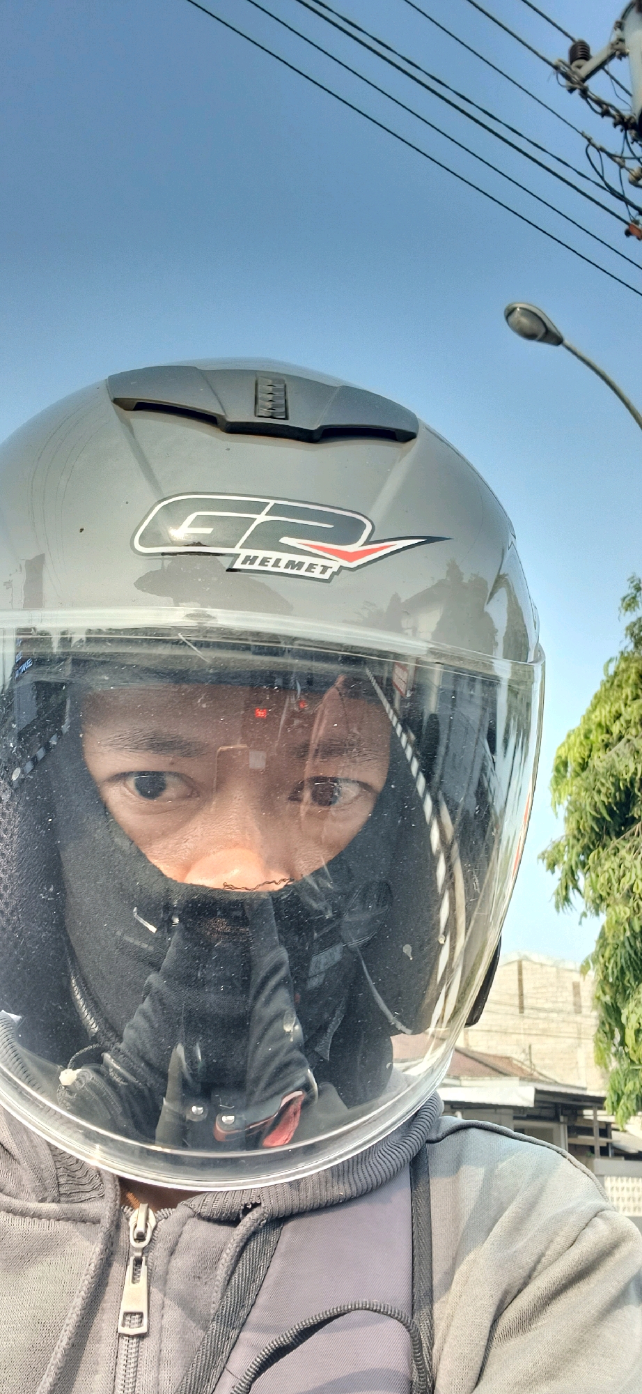 A close-up, first-person perspective selfie taken by an individual wearing a gray motorcycle helmet with a clear visor, a black balaclava covering their face from the nose down, and a gray hooded jacket. The individual's eyes and eyebrows are visible through the visor. The helmet has a "G2 HELMET" logo. The background shows a clear blue sky, overhead power lines, a street lamp, and some trees and buildings, suggesting an outdoor urban or suburban environment. The overall scene appears to be in Kranggan, Indonesia, based on the context provided. The lighting suggests it is daytime.