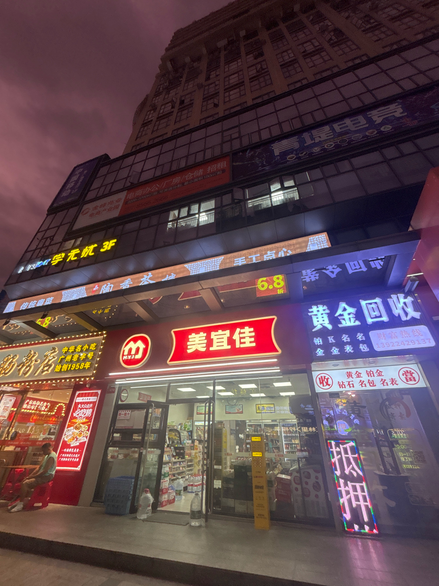 This image captures a nighttime street scene in Nanhai District, China, dominated by a tall, modern building with multiple illuminated signs. The sky is a dusky purple, suggesting evening twilight. The ground level features several businesses.
To the left, a restaurant or food establishment is prominent with a large, illuminated sign reading "肠鹅启" and further text in Chinese. A person is seated on a red stool outside this establishment.
Adjacent to it, a store with a red and white sign that reads "美宜佳" is visible. The entrance to this store is open, revealing shelves stocked with products.
To the right of "美宜佳", there's a brightly lit sign for "黄金回收" (Gold Recycling). Below this, another sign displays icons and text related to gold, diamonds, and bags. Further to the right, a vertical, illuminated LED sign displays Chinese characters in vibrant colors.
The building above the storefronts has several floors with numerous windows. The lower floors of the building feature larger glass windows and more illuminated signage. One sign reads "青海电竞" (Qinghai E-sports), and another, in yellow, reads "学无忧 3F" (Learn Without Worry 3F). Other signs advertising various services and businesses are also present. The overall impression is a busy commercial area at night.