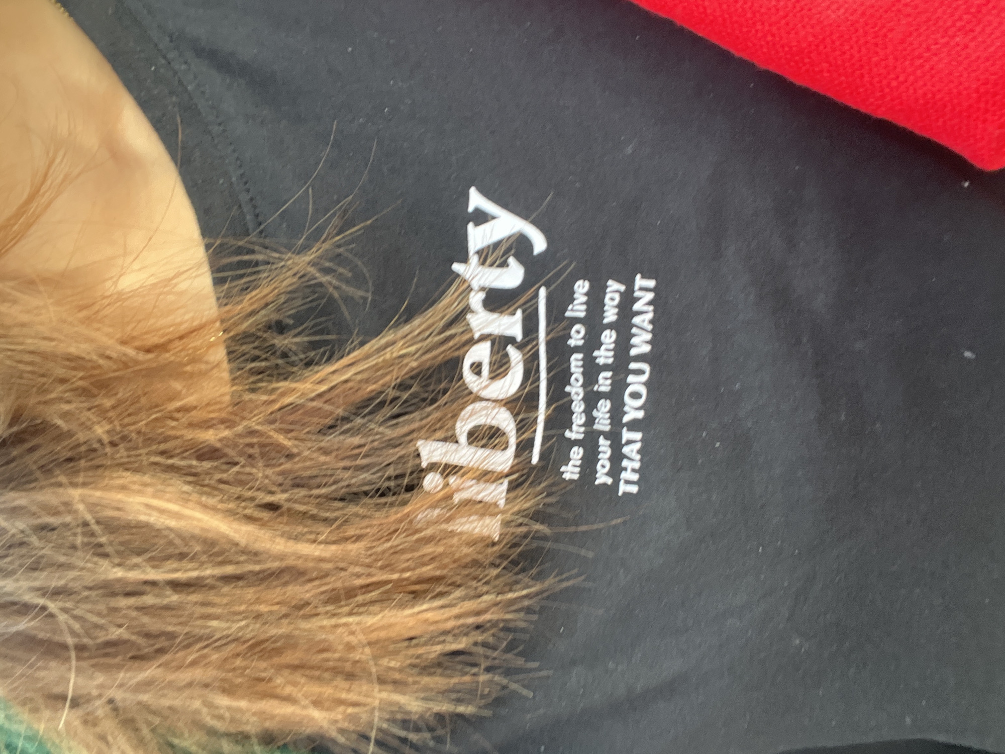 A person's shoulder and upper chest are visible in the image, against a black t-shirt with the text "liberty the freedom to live your life in the way THAT YOU WANT" printed in white. The person's blondish-brown hair is spread across the shirt. A red object appears in the upper right corner of the frame. It is not possible to determine the location in Davao City, Philippines, from the given image.