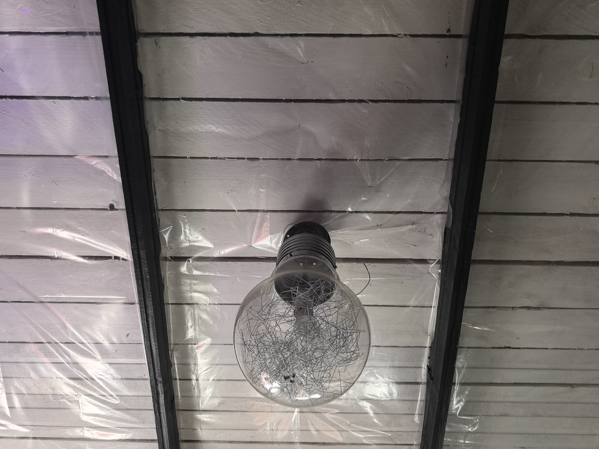 The image displays an overhead view of a ceiling or angled wall section located in Noviy, Russia. A large, spherical clear glass light fixture is centrally mounted on a surface composed of horizontally arranged, light-colored wooden planks, likely painted white or pale grey. The interior of the glass sphere contains a dense, chaotic arrangement of thin, bent metallic wires. The fixture's base is dark, with visible horizontal ridges, and is directly attached to the wooden structure. The entire overhead surface, including the wooden planks and parts of the light fixture's base, is covered by a layer of clear, wrinkled plastic sheeting, which reflects ambient light. Two dark, structural beams or dividers run diagonally across the frame, partially obscured by the plastic sheeting. No individuals or active subjects are present in the scene.