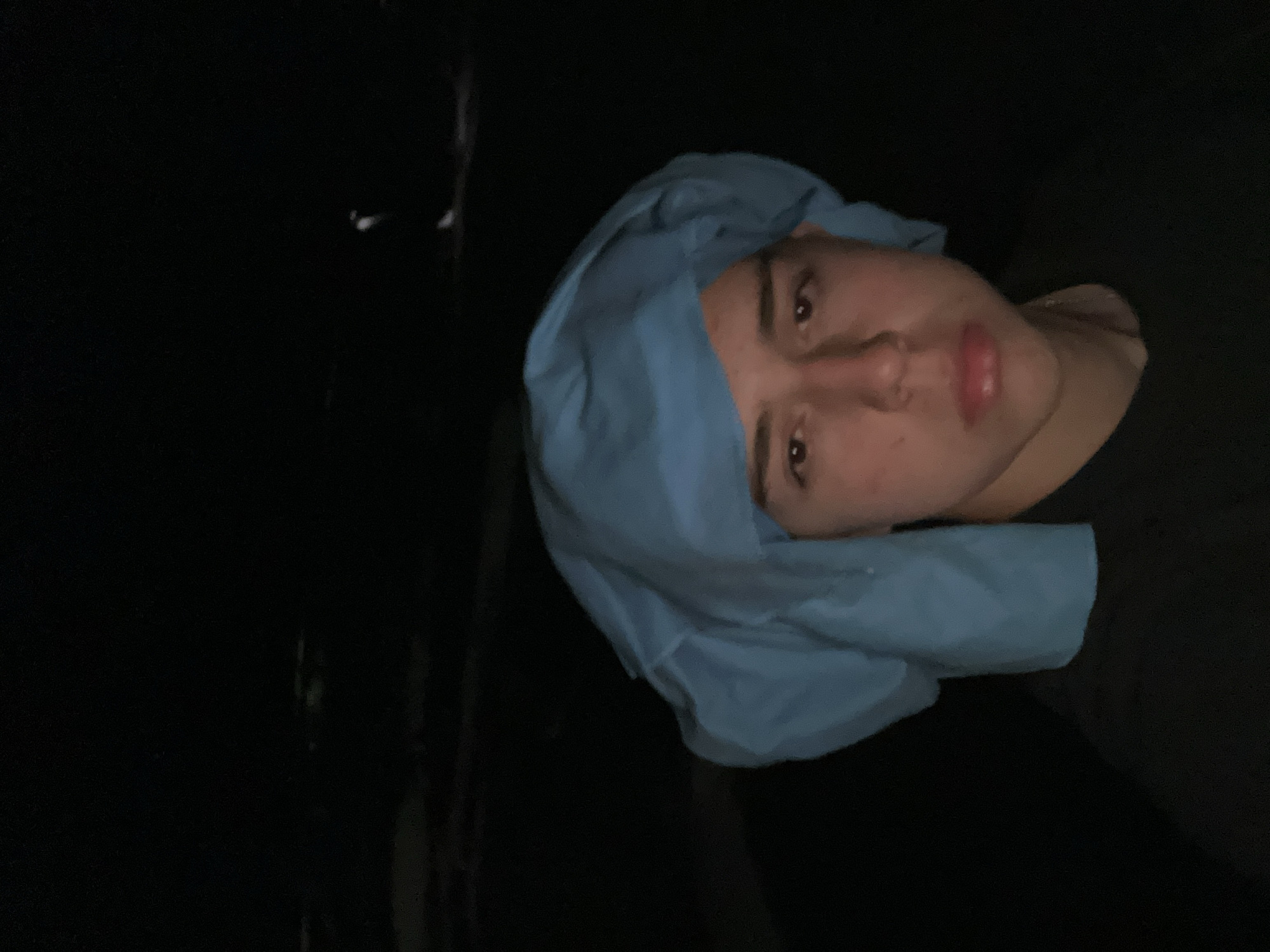 A dimly lit photograph shows a person wearing a light blue covering on their head. The subject is positioned in the right portion of the frame, with a dark, undifferentiated background filling the left side. The individual's face is visible, with eyes open and lips slightly parted. A small portion of the person's dark clothing is also visible, situated beneath the subject's face. The image is oriented so the subjects head is toward the right, and the background takes up the space that would be below. The Iamatikes Piges, Greece location is not confirmed or denied.