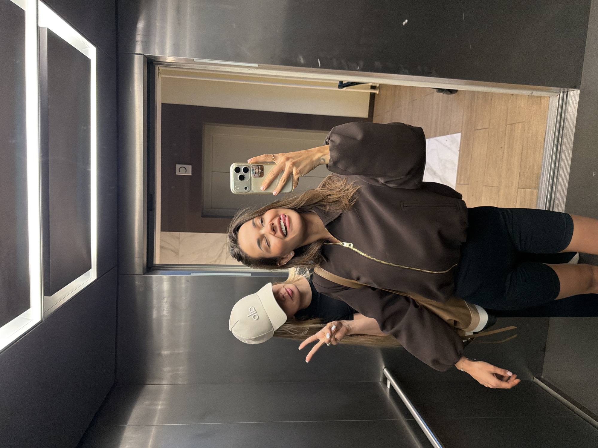 In an elevator in Moscow, Russia, two young women are taking a selfie. The woman in the foreground is holding a smartphone in her right hand, capturing the reflection in the elevator's mirrored walls. She is wearing a brown zip-up jacket and black shorts, with her tongue sticking out and eyes closed. Behind her, another woman wearing a tan baseball cap with "alo" logo is peeking over her shoulder, making a peace sign with her left hand. The elevator has a stainless steel interior and fluorescent lighting.
