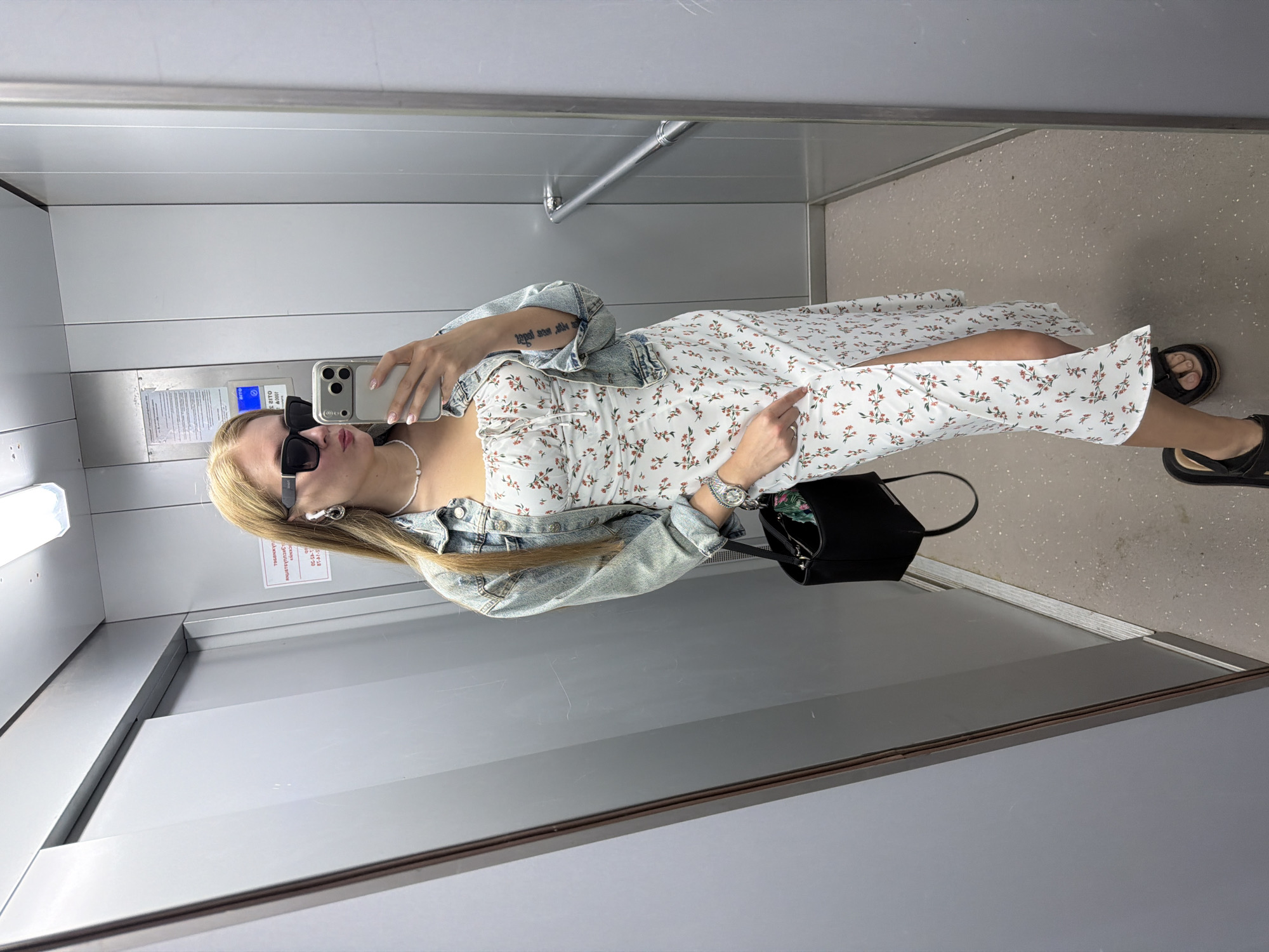 A woman is depicted taking a full-body mirror selfie inside an elevator, likely in Chaikovsky, Russia, as specified by the prompt. She has long, straight blonde hair and is wearing large black sunglasses, obscuring her eyes. Her lips are slightly pursed in a selfie pose. She is dressed in a light blue, unbuttoned denim jacket over a white midi-length sundress with a delicate pattern of small reddish-orange flowers and green leaves. The dress features thin straps, a tie-front detail at the bust, and a high slit on the left side, revealing her upper thigh. Her footwear consists of black platform sandals.
Accessories include a white, pearl-like choker necklace, silver-toned hoop earrings, and white wireless earbuds in her ears. A metallic wristwatch with a large, round face is visible on her left wrist, and a ring adorns her right ring finger. She holds a light-colored smartphone (consistent with an iPhone model) in her right hand, positioned to capture her reflection. A black rectangular handbag with a short strap hangs from her left hand, which is resting by her side, her index finger extended near the dress slit. A tattoo of text is visible on the inside of her left bicep/tricep.
The elevator interior features light grey panelled walls, a rectangular fluorescent light fixture on the ceiling, and a light, speckled grey floor. A silver-colored metal handrail is affixed to the right wall (visible in reflection). On the wall reflected behind the woman, a white sign with red Cyrillic text is visible. Key phrases on the sign include "УВАЖАЕМЫЕ ЖИТЕЛИ!" (Dear Residents!), "РЕМОНТ" (Repair), and a date "26.05.2023" (May 26, 2023), along with times "С 09-00 ДО 17-00" (From 09:00 to 17:00).