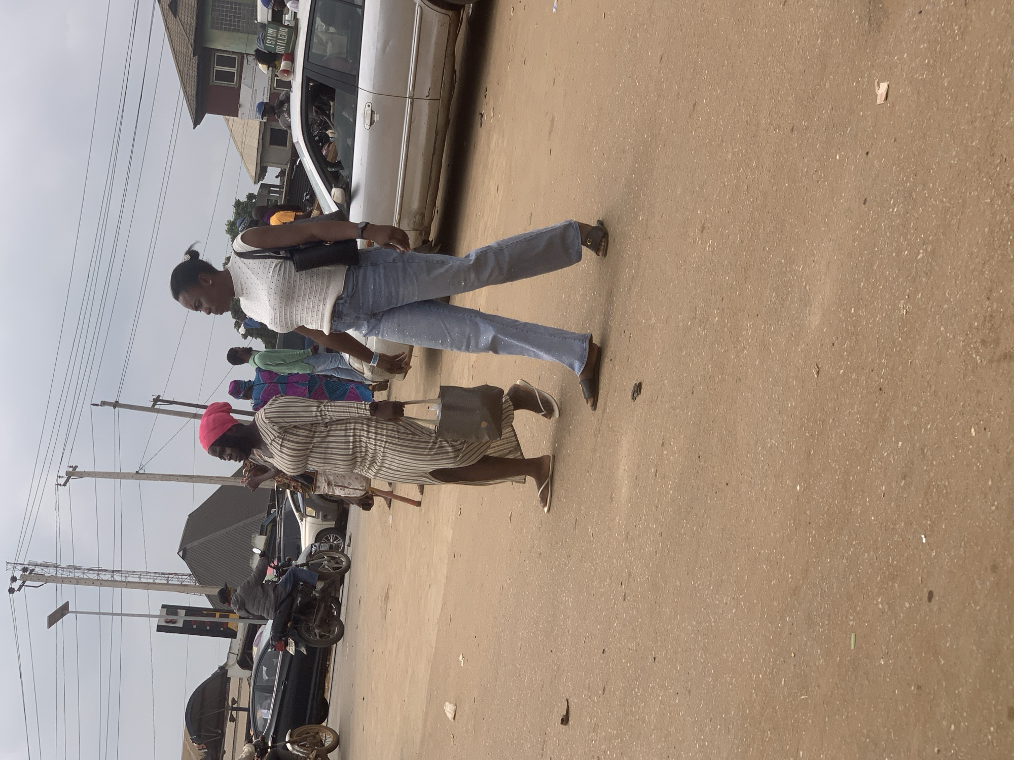 In the city of Shagamu, Nigeria, two women are walking across a dusty, unpaved road. The woman on the left is wearing a striped dress and a pink headscarf, carrying a dark handbag. The woman on the right is wearing a white, high-necked, short-sleeved top and light blue jeans, also carrying a dark handbag. They appear to be walking away from the viewer. In the background, there are various vehicles, including cars and motorcycles, as well as utility poles and buildings. The sky is overcast.