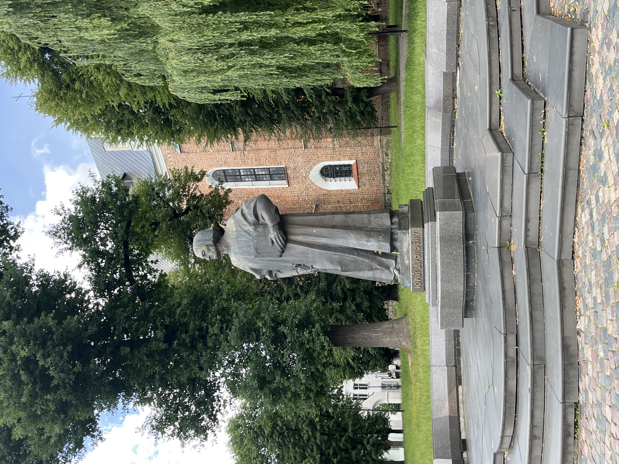 In Drohobych, Ukraine, a bronze statue of Yuriy Drohobych stands prominently on a tiered, dark stone pedestal in front of a red brick building with arched windows. The statue depicts a man in a long robe, holding a book. He is positioned centrally in the frame, facing to the viewer's left. The surrounding environment includes lush green grass, tall trees with dense foliage, and a cloudy sky. To the left of the statue, a section of a white building is visible. The ground in the foreground is paved with cobblestones. The overall scene captures an outdoor public space with historical and architectural elements.