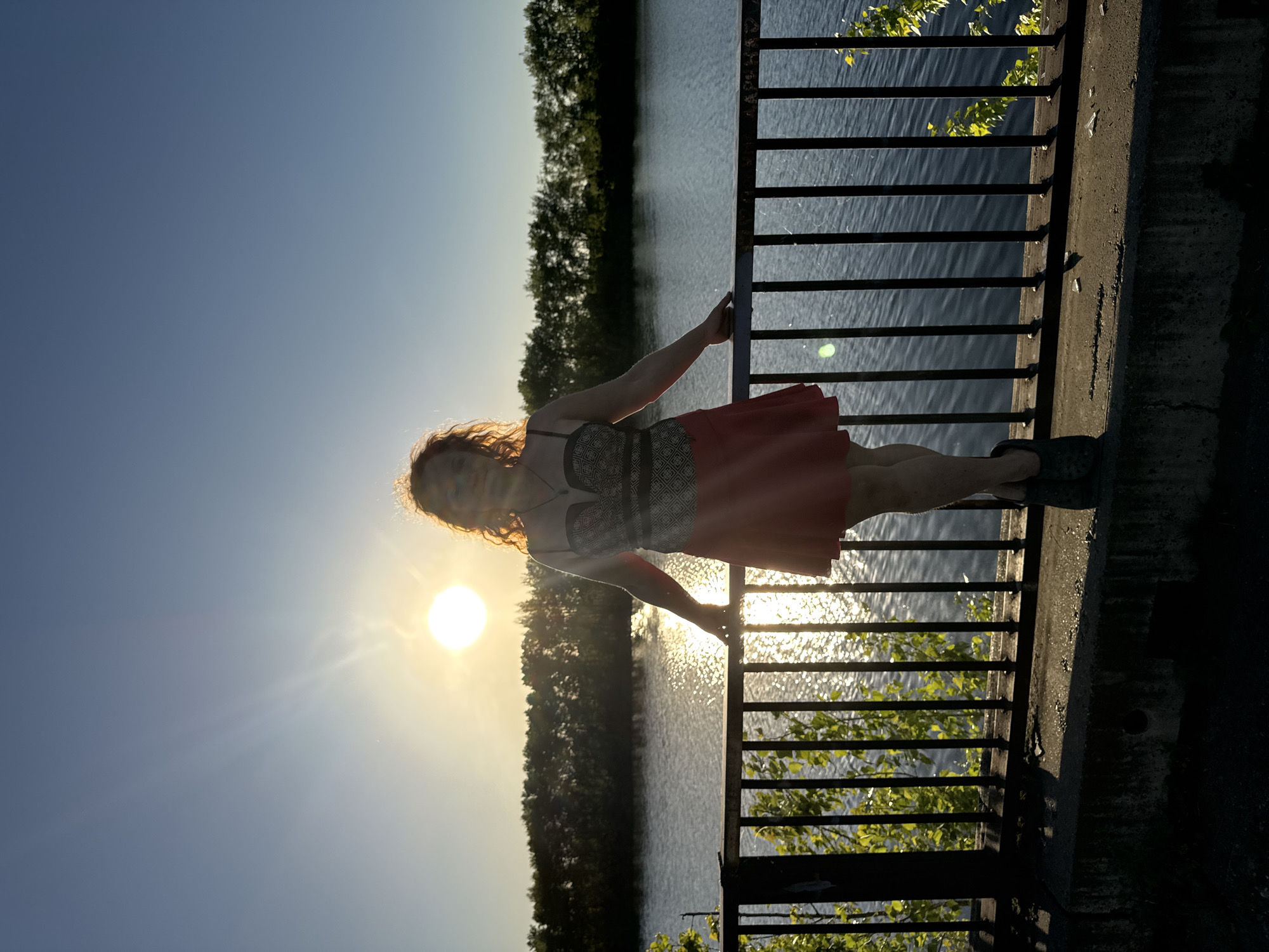 A vertical, medium shot depicts a person standing on a concrete structure overlooking water and trees. The person is facing the camera, holding onto a dark, horizontal railing. They are wearing a red skirt and a gray top. Behind them, the setting sun reflects brightly on the water. The opposite bank of the water feature is lined with trees. The sky is pale blue, with a bright sun casting a strong glare. The location is likely a bridge or overlook in Chaikovsky, Russia.