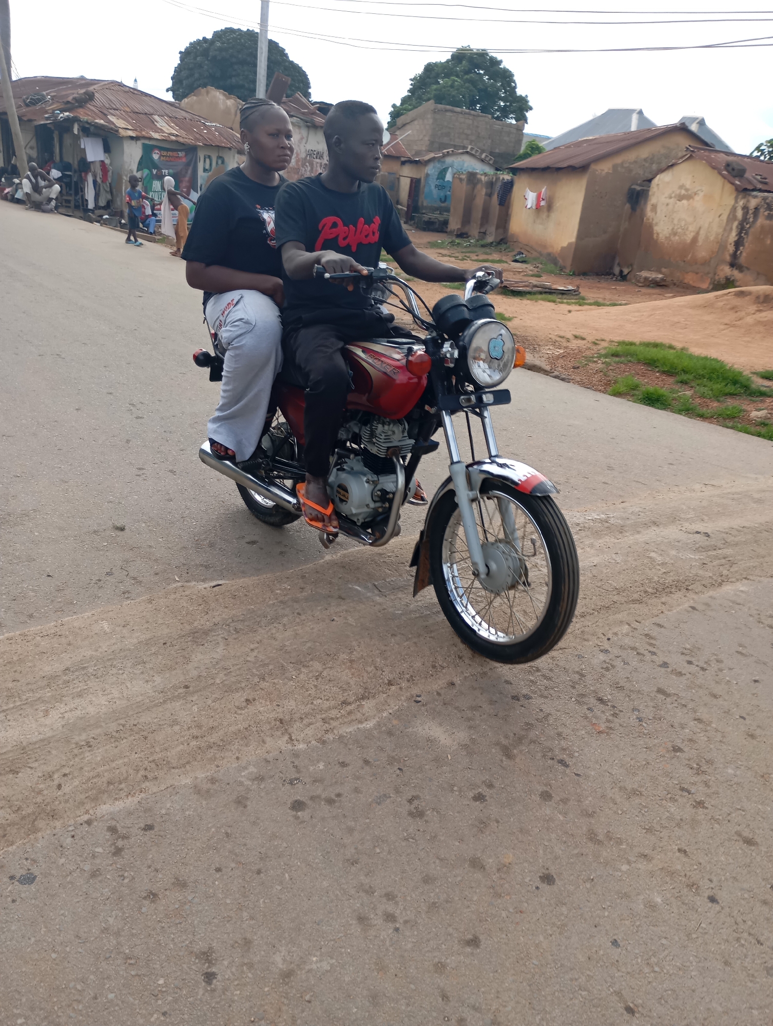 In Shendam, Nigeria, two individuals are riding a red and silver motorcycle on a paved road. The person in the front, a male wearing a black t-shirt with red lettering that reads "Perfect," is driving. The person behind him, a female wearing a black t-shirt and light grey pants, is a passenger. Both are looking forward. The motorcycle has a circular headlight and a chrome exhaust pipe. The road surface appears worn and dusty, with some oil stains visible. In the background, there are buildings with corrugated iron roofs and a few trees. Other people and a young child can be seen in the distance, near some of the buildings. The overall setting appears to be a residential or commercial area in a town.