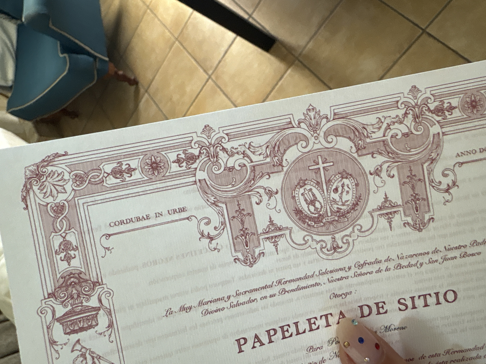 Here is a concise description of the image:
The image shows a close-up of a decorative certificate or document, likely religious in nature. The text on the document includes phrases like "CORDUBAE IN URBE", which indicates a connection to the city of Córdoba, Spain. Other phrases mention "Hermandad Salesiana" (Salesian Brotherhood) and "Sacramental Kermandad." The term "PAPELETA DE SITIO" is prominently displayed, suggesting it might be a document related to membership or participation in a religious event or organization. The certificate has an ornate border design with religious symbols such as crosses. Part of a manicured finger with nail art is visible on the lower part. The image is taken indoors, with part of a blue chair and tiles visible in the background, suggesting the document is being held or placed in a room with tiled floors.