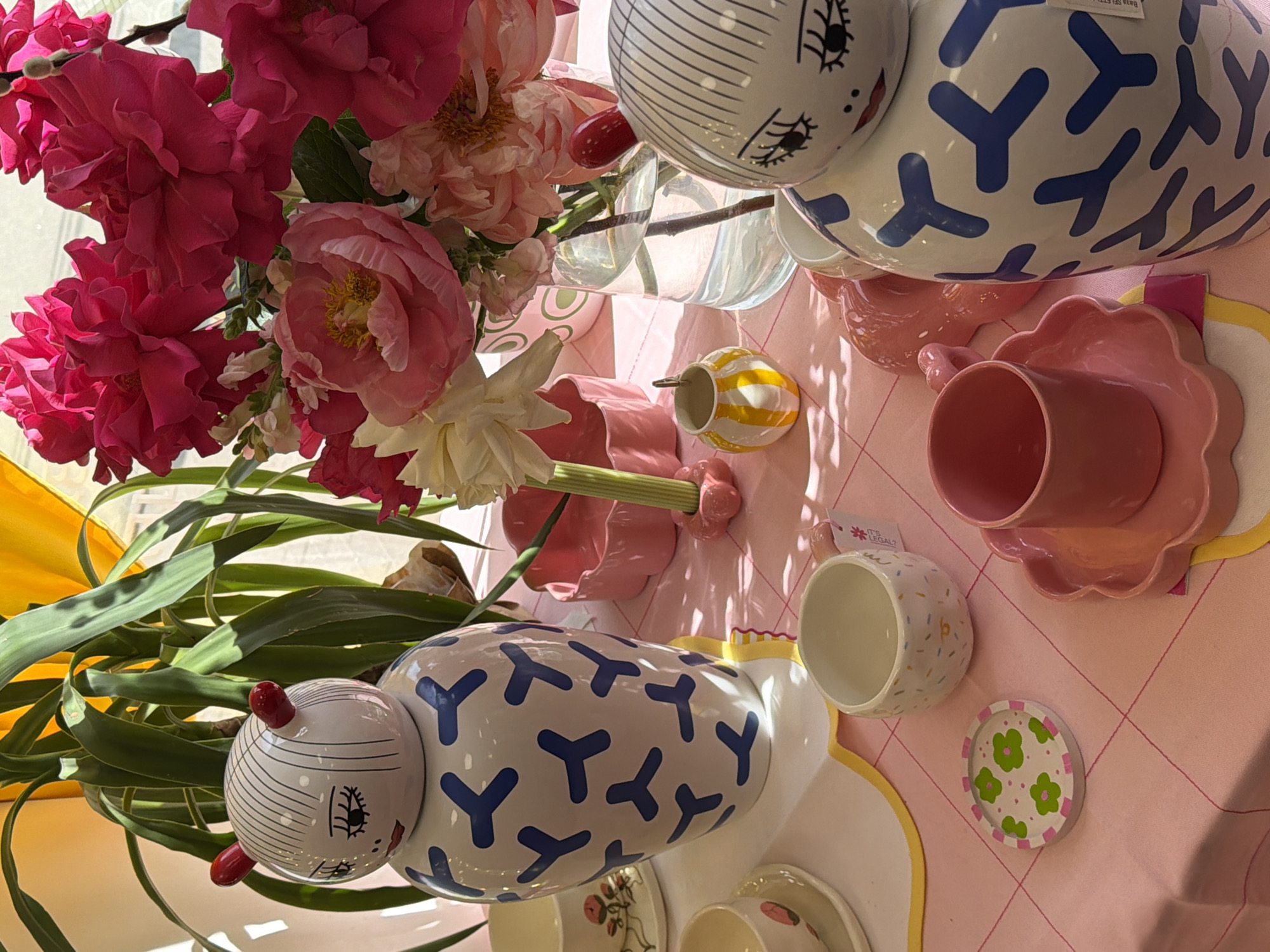 An overhead shot captures a brightly lit table display featuring a vibrant arrangement of flowers and an assortment of decorative ceramic tableware. No people are visible.
The central focus is a large bouquet dominated by lush pink and coral peonies, interspersed with delicate white flowers and vibrant green foliage. Bright light from the upper left casts soft shadows across the scene.
Arranged on a pink tablecloth with a subtle red diagonal grid pattern are various ceramic items. Two prominent white, elongated ceramic figures resembling stylized caterpillars or snails are visible. Each features a cartoon-like face with closed eyes, long eyelashes, and red lips or cheeks, topped with red antennae-like protrusions. Their bodies are adorned with a repeating blue Y-shaped pattern. One figure is positioned at the top right, and the other at the bottom center.
Around these figures are several smaller ceramic pieces: a small, round yellow cup with white stripes; a pink cup and matching saucer with a scalloped edge; a white cup patterned with colorful sprinkles; and a small, round white coaster or dish decorated with green clover-like flowers. Other partially visible ceramic dishes and cups are seen towards the bottom of the frame.
A small white tag, partially visible and attached to a cup, bears the text "IT'S LEGAL!". Another tag on one of the larger ceramic figures shows unreadable text and "Baza SST23...". The arrangement suggests a whimsical and cheerful aesthetic, possibly a display of home goods or a decorated table for an event.