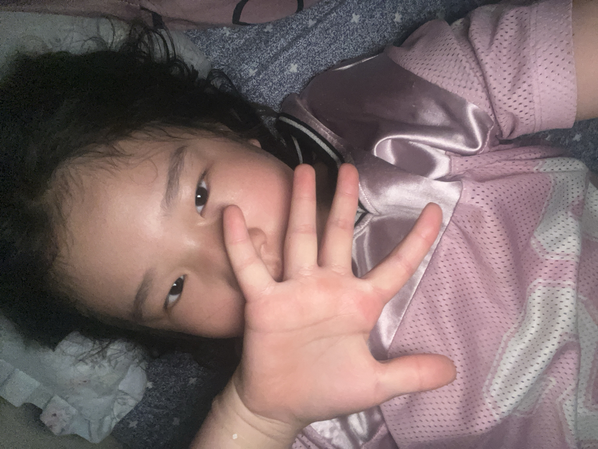 A young child lies on a bed, their head resting on a blue patterned sheet. They wear a pink jersey with white trim and a satin-like collar. Their face is partially obscured by their hand, fingers outstretched towards the camera. The background includes sections of the bed sheet and a white ruffled fabric.