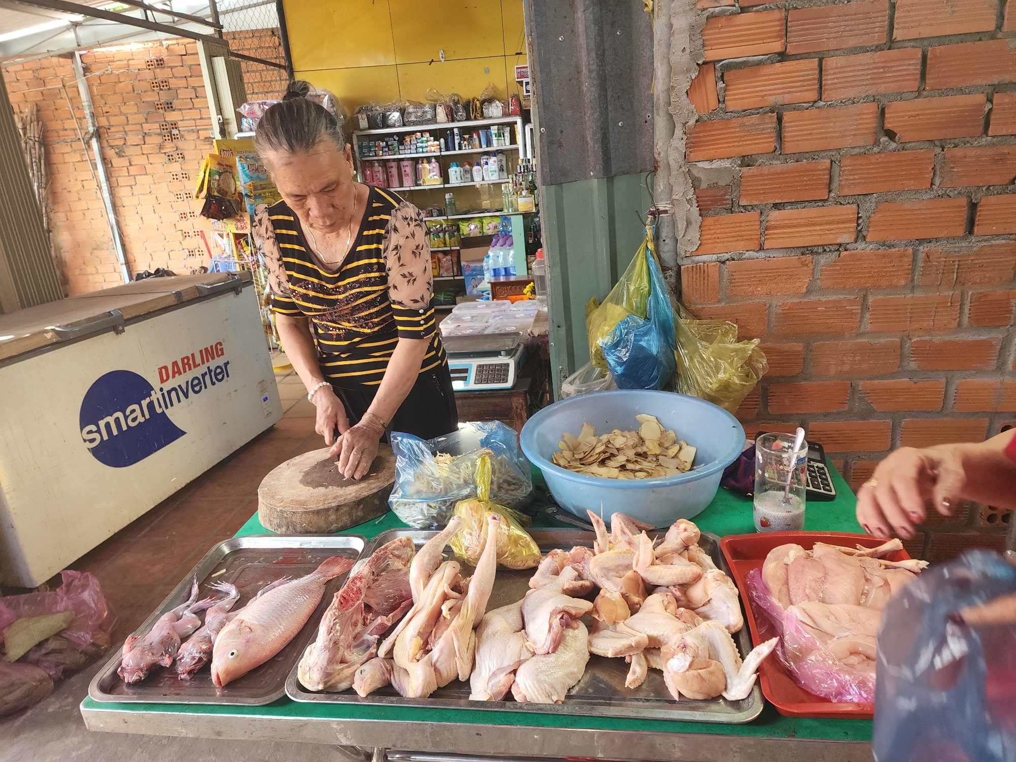 In Phu Thuy Ward, Vietnam, a woman is seen preparing raw chicken at a market stall. She is standing behind a counter laden with various food items, including whole chickens, chicken parts, and fish. A cutting board and knife are positioned in front of her, indicating she is actively engaged in processing the meat. The stall is set up outdoors, with a brick wall visible in the background and shelves stocked with goods. A large refrigeration unit is present on the left side of the frame, and a scale is visible on the counter. The scene suggests a typical day at a local Vietnamese market, with an emphasis on fresh produce and meats.