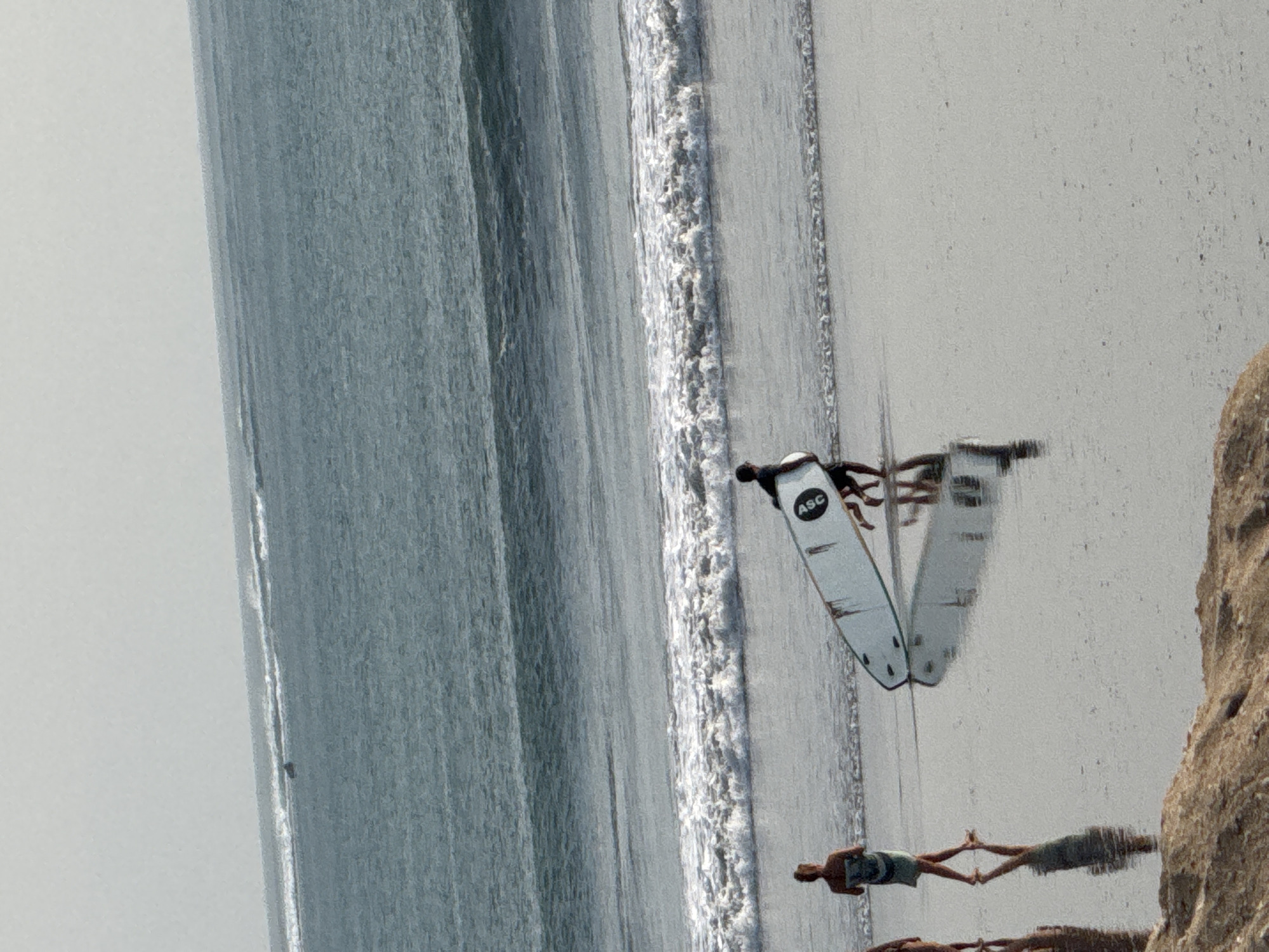 The image depicts a beach scene in Kuta, Indonesia. Three individuals are visible on the wet sand near the water's edge. In the foreground, a person is walking away from the camera, carrying a surfboard. The surfboard is white and has black fin placements. To the left, another person is walking towards the camera. Further down the beach, a third individual is walking away from the camera, also carrying a surfboard. The ocean is in the background, with waves breaking on the shore. The sky is overcast. The sand is wet and reflective, indicating recent tide activity.