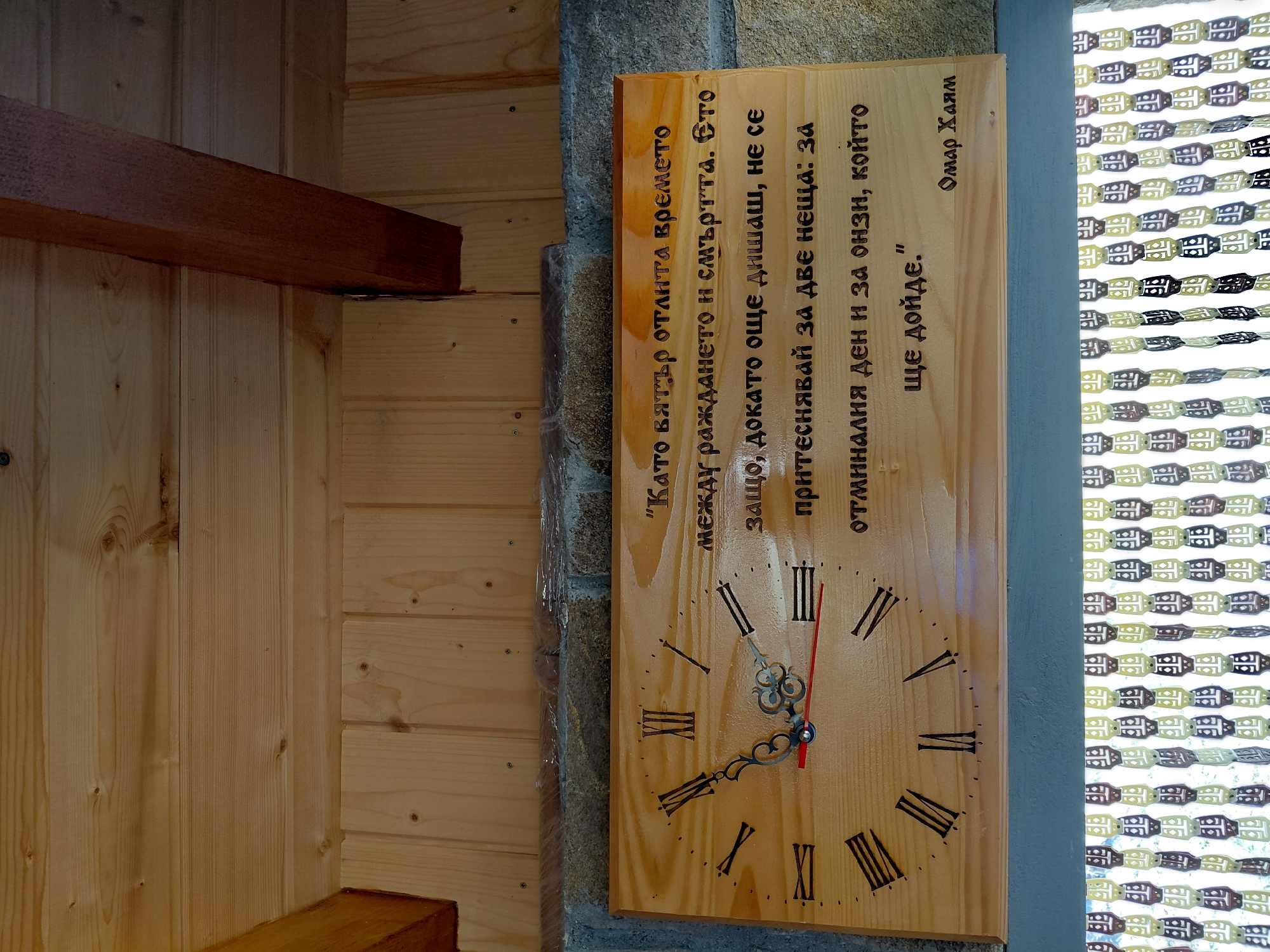 The image shows a wooden clock face with Roman numerals and text etched into it, hung on a stone wall with a wooden ceiling above. The text is in Bulgarian and appears to be a quote. The clock hands are visible, with a red second hand. Below the clock, there is a beaded curtain. The environment suggests an indoor or covered outdoor space, possibly a home or a rustic establishment. The location is specified as Dryanovets, Bulgaria.