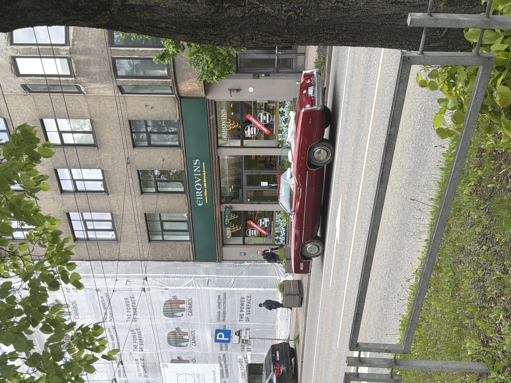 This media file captures an urban street scene in Riga, Latvia, on what appears to be an overcast daytime. The image is oriented sideways, but described as if rotated upright.
The prominent subject is a red vintage convertible car, with chrome details, parked on the right side of the street, parallel to the sidewalk. Further down the street, a black modern car is partially visible, also parked. The street features asphalt pavement with white painted lines.
To the left of the street stands a multi-story building. Its ground floor houses a shop with a green facade and large windows. The shop is clearly identified by multiple signs reading "EIROVINS" and "VINU LIELVEIKALS" (Wine Supermarket). Additional text on the shop windows includes "ATVERTS KATRU DIENU" (Open every day) and "www.eirovins.lv". Above the shop, the building has several floors with numerous rectangular windows.
A significant portion of the building to the left is covered by a white construction tarp, indicating renovation or construction work. Text visible on the tarp includes "CAPAROL", "KRĀSAS APMETUMI FASĀDES SILTINĀŠANA" (Paints, Plasters, Facade Insulation), and "THE POWER OF SURFACE".
Two men are visible on the sidewalk; one stands near the EIROVINS shop entrance, and another is further down, near a planter on the sidewalk, partially obscured by the construction tarp. Green trees and foliage are present in the foreground and along the street, suggesting mild weather and a spring or summer season. Metal barriers or fences are also visible bordering the street and green areas. A blue parking sign with a white "P" and "10 min" indicates parking regulations. The scene depicts a typical city environment with commercial activity and ongoing development.