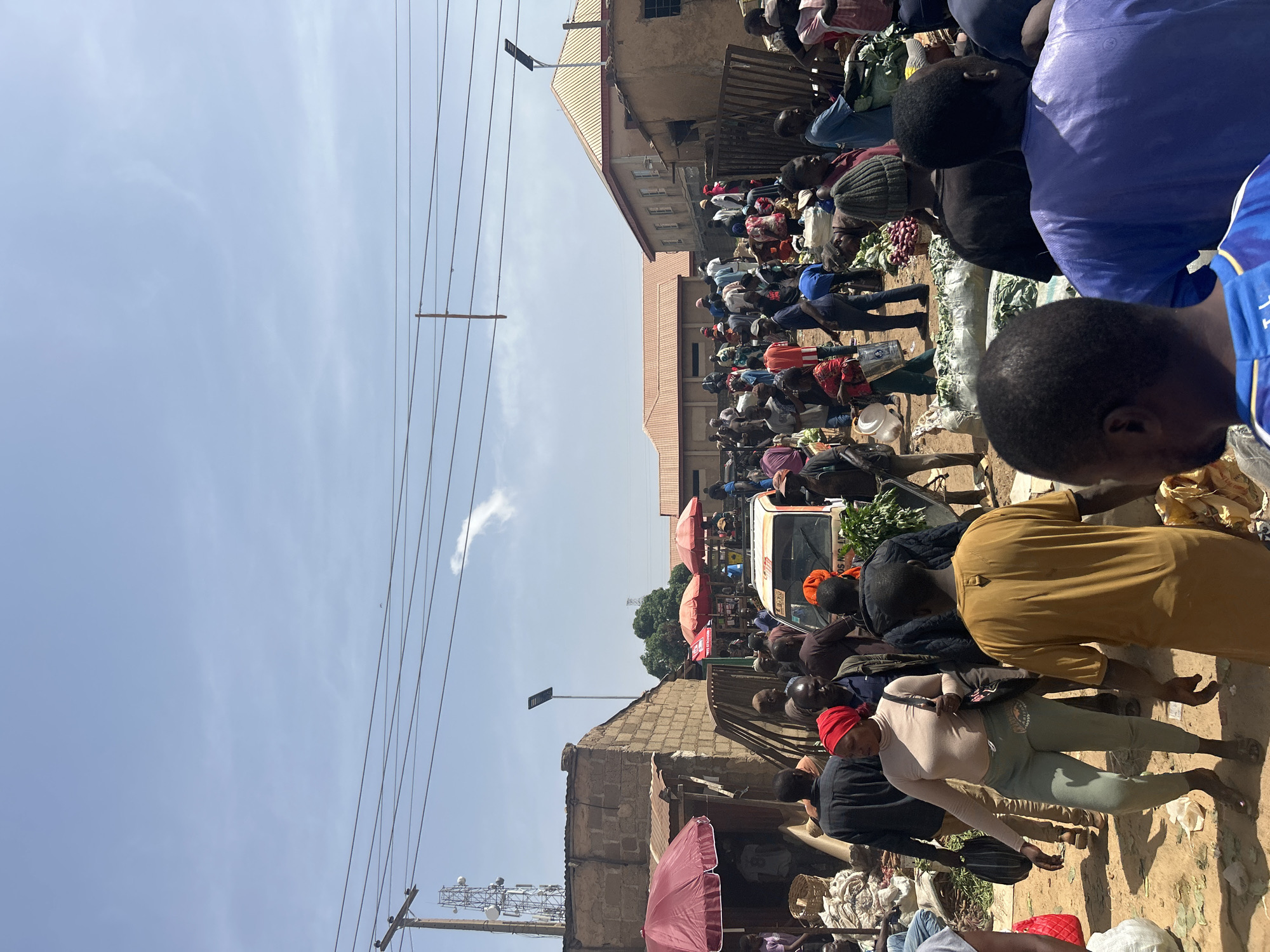 The image captures a crowded street scene in Bukuru, Nigeria. Numerous people are visible, many gathered closely together, suggesting a market or busy commercial area. Various individuals are walking, standing, and appearing to engage in transactions. Goods are displayed, including what appears to be produce. A white van is parked in the middle of the street. There are red umbrellas. Buildings of varying heights and construction types line the street. Power lines are visible against a light blue sky. The ground appears unpaved or dusty.