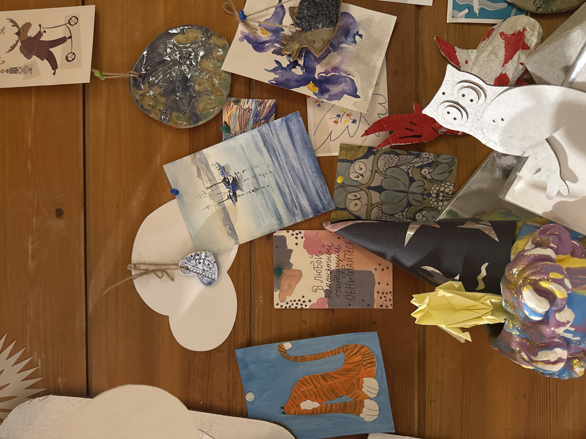 This media file showcases a vibrant collection of diverse handmade artworks and crafts, predominantly pinned or arranged on a warm, vertical wooden surface, likely a wall or board. The display suggests a personal or communal exhibit of creative expression.
Among the subjects are a whimsical, cartoon-style drawing of a moose-like creature on a wheeled contraption, located at the top left. Below it, a white, spiky sunburst paper cut-out is partially visible. A circular, glazed ceramic piece with abstract blue and brown textures, resembling a stylized globe or celestial body, is prominently featured.
Several watercolor paintings are visible: one with broad, abstract purple and blue strokes, possibly depicting irises, and another with vertical blue brushstrokes and reflections, suggesting a water scene. There are various paper cut-outs, including a white stylized animal head with large concentric eyes and a red flame-like mouth, and other abstract red and white paper shapes. A small, white, heart-shaped ceramic piece is attached with twine to a larger white cloud-shaped paper cut-out.
Other items include a black fabric piece adorned with silver star cut-outs, a yellow-green origami frog, and a brightly painted, multicolored ceramic sculpture with glitter accents. A painting of the back half of an orange and black striped tiger on a blue background is situated towards the bottom. A piece of patterned paper featuring stylized owls can also be seen.
A rectangular card with an abstract painted background is centrally located and contains Russian text: "В любой непонятной ситуации ОБНИМАЙТЕСЬ," which translates to "In any incomprehensible situation HUG." The overall arrangement conveys a sense of playful creativity and personal touch, aligning with the provided context of Moscow, Russia, where such a display could be found in a home or art space.