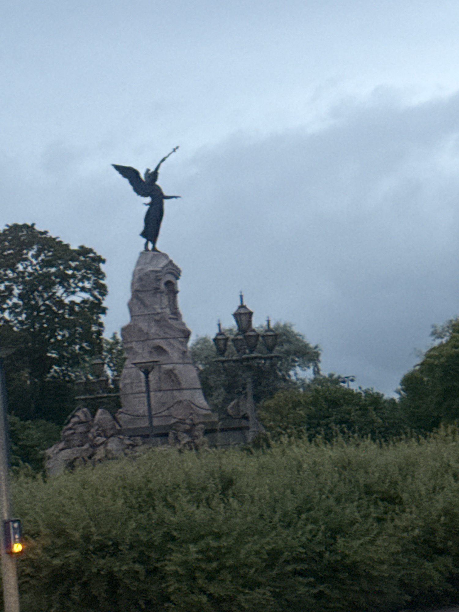 The image shows a monument with a winged figure at the top, standing on a stone structure. The figure appears to be an angel or victory statue, holding a spear or staff. The monument is surrounded by trees and greenery. In the background, there is a cloudy sky and more trees. A streetlamp is visible in the foreground. The monument is located in Kadriorg Park, Tallinn, Estonia, and is known as the Monument to the Victory of the War of Independence.