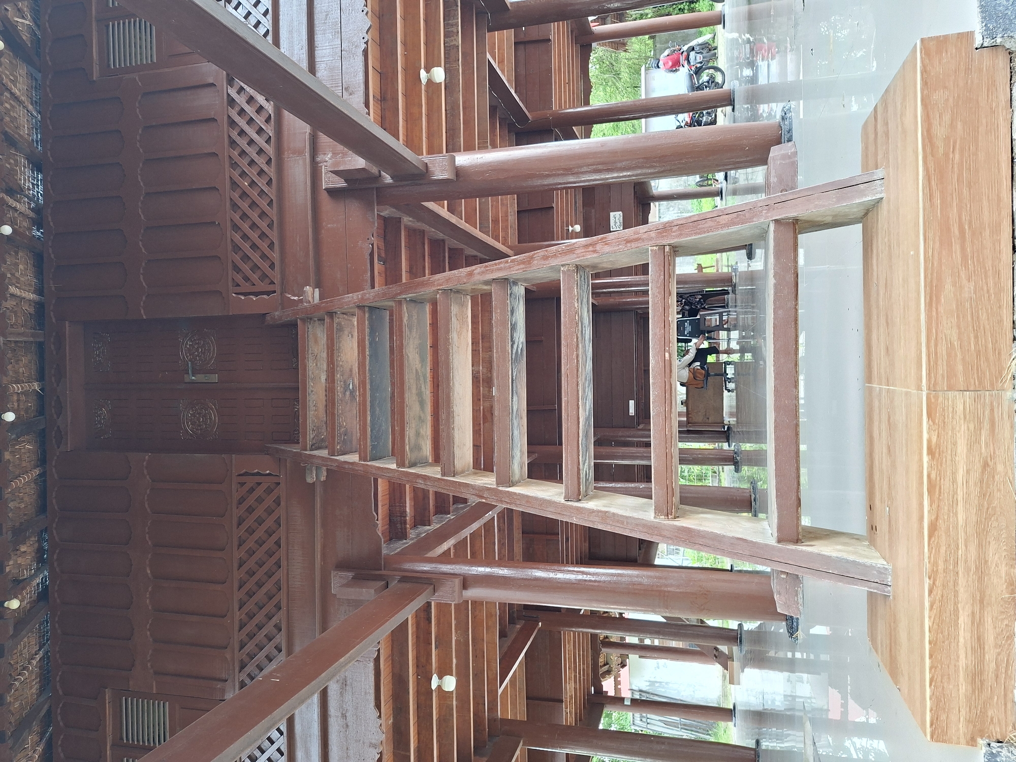 The image captures a perspective view of a wooden structure, possibly a traditional building or house, in what appears to be Lhokseumawe, Indonesia. The primary focus is the intricate woodwork of the building, showcasing a lattice design and decorative carvings. Visible are wooden beams and posts supporting the structure. A set of stairs are visible. Outside the building, blurred glimpses of the outdoor environment suggest a lush, green landscape with vegetation. A parked scooter. A polished, reflective surface, possibly a floor, mirrors the building's framework and the surroundings, enhancing the perspective and depth of the image. A camera crew in the reflection on the surface.