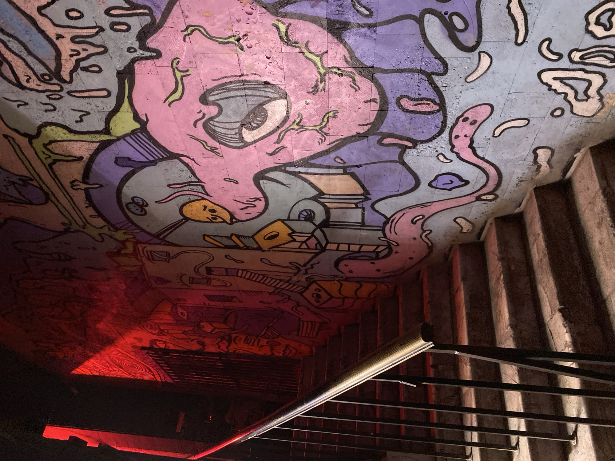 The image depicts a staircase leading downwards, illuminated by a red light source at the bottom, possibly a bar or club. Above the staircase, a graffiti-covered ceiling features cartoonish figures and abstract shapes in shades of pink, purple, blue, and grey. The staircase is constructed of concrete and includes a metal railing. The location is in Moscow, Russia, based on the graffiti style which is unique to that location.