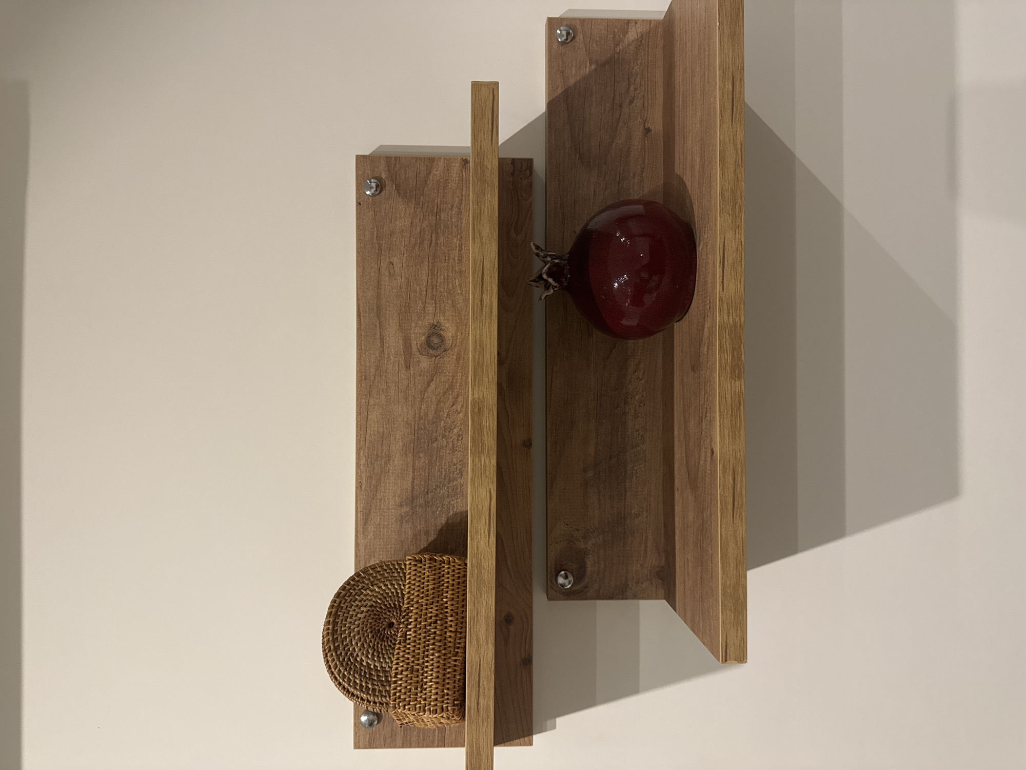 A vertically oriented, eye-level shot features a decorative wooden wall hanging against a plain, light-colored wall. The hanging consists of two vertical wooden panels of different lengths, each affixed to the wall with two visible metal screws. A slender wooden strip is positioned between the two panels. A round, woven basket sits near the bottom of the left panel, while a glossy, deep red pomegranate rests on the right panel. The right panel casts a distinct shadow on the wall. The location is unknown.