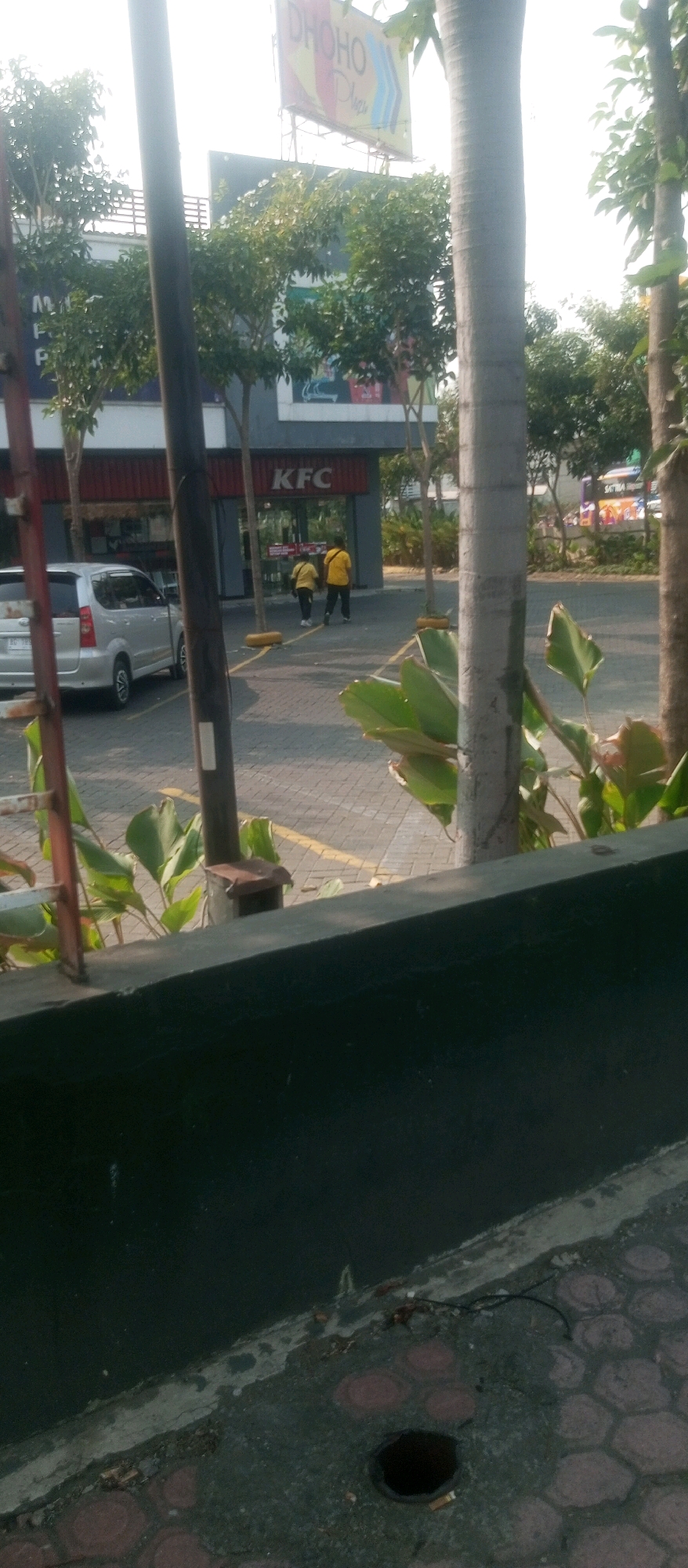 In Kediri City, Indonesia, a silver Toyota Avanza is parked in front of a KFC establishment. Two individuals wearing yellow shirts are walking away from the KFC. The scene is set outdoors, with trees and a paved area visible. A large sign, partially obscured, reads "DHOHO Plaza." In the foreground, a dark green planter box is present, and beyond it, a patterned paved surface with a circular opening is observed.